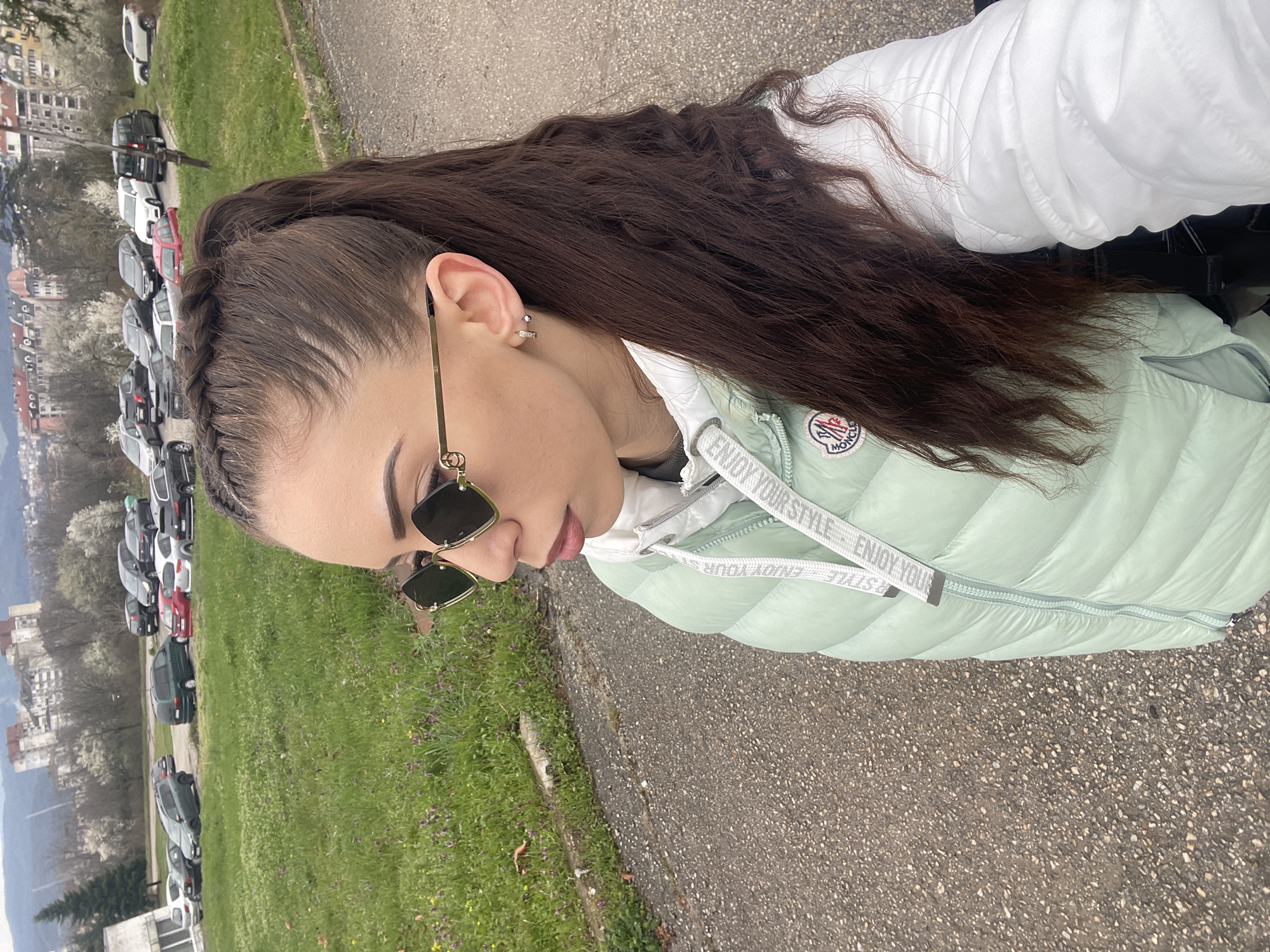 A young woman stands on a paved road. The background reveals a cityscape and a row of parked cars, likely the city of Veliko Tarnovo, Bulgaria. The woman is wearing a light green puffer jacket over a white hoodie, and sunglasses. Her hair is styled with a braid along the hairline. The pavement is textured and gray. A patch of green grass and vegetation are visible on the left, beyond which is the cityscape.