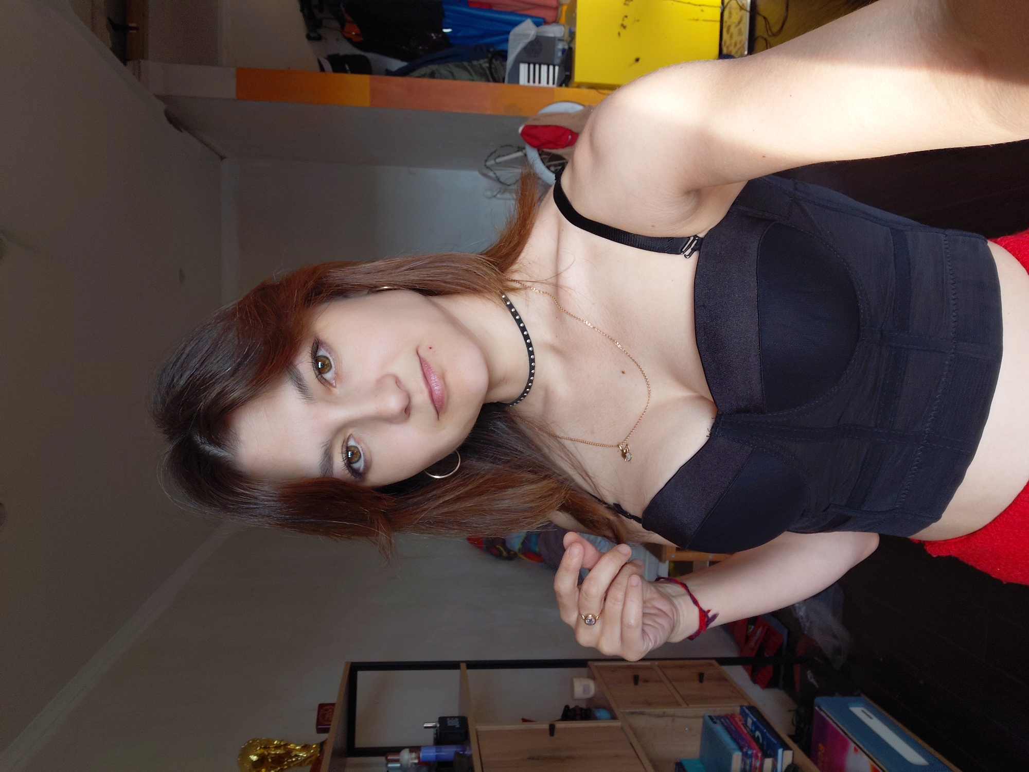 In a dimly lit room, a young woman with medium-length auburn hair and brown eyes looks directly at the camera. She is wearing a black corset-style top and red bottoms. She has a black choker with studs around her neck and a delicate gold chain necklace with a small pendant. A gold ring adorns her right index finger, and a red bracelet is visible on her left wrist.
The background reveals a room with shelves filled with various items, including bottles, books, and decorative objects. A trophy is visible on the top shelf to the left. To the right, a bright yellow surface is present, with clothing items and what appears to be an accordion-like object in the background. The lighting in the room is uneven, with a strong light source casting shadows. The overall impression is that of a personal space, possibly a bedroom or living area. The city of Magnitogorsk, Russia, is not visually indicated in the image.