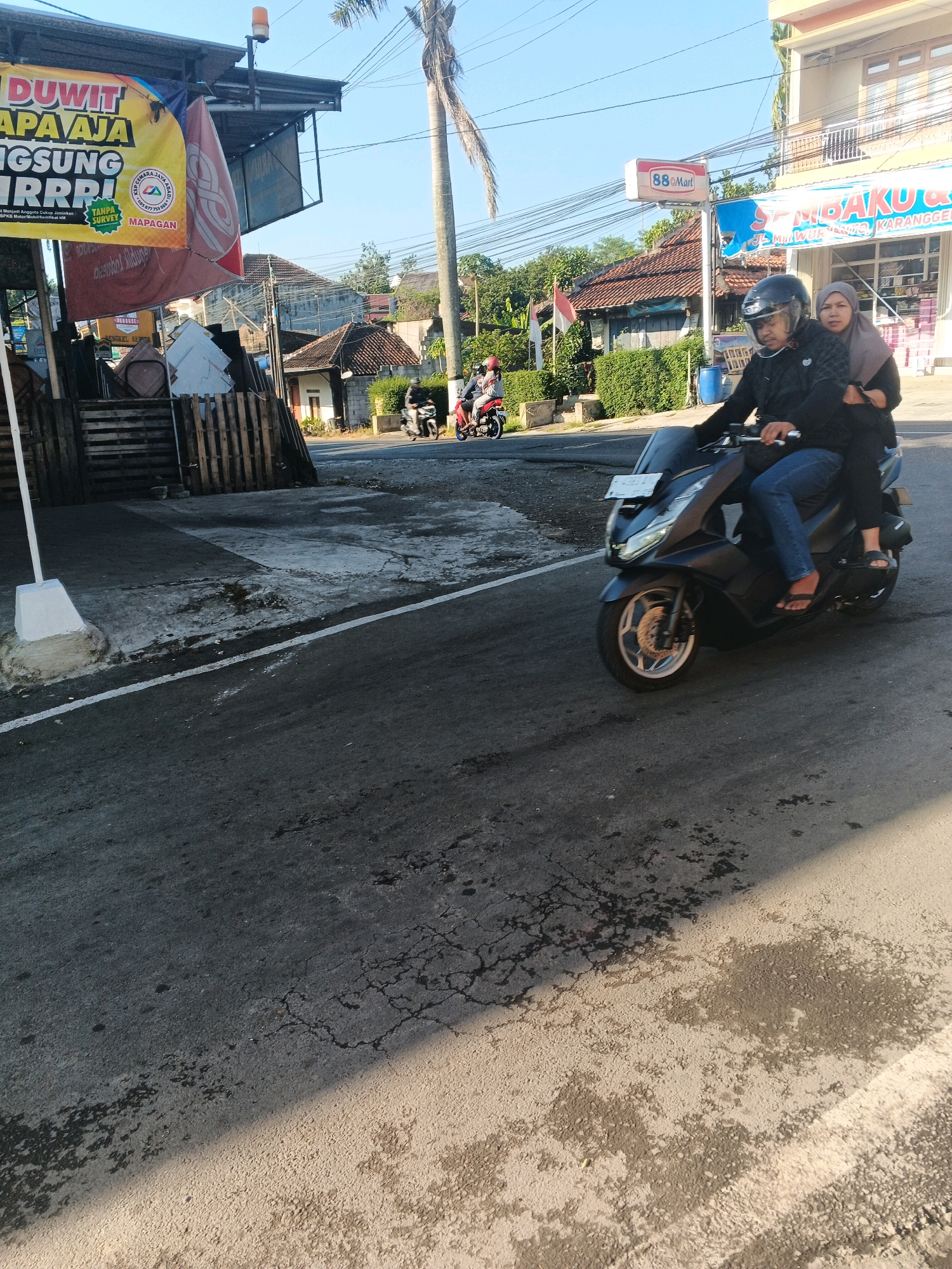 In Semarang, Indonesia, a man wearing a black jacket and blue jeans rides a dark-colored scooter. He is wearing a black helmet. A woman wearing a grey hijab and black clothes sits behind him, holding onto him. They are traveling on a paved road with some cracks and dark stains. In the background, there are buildings, trees, and a blue sky. A sign for "88 Mart" is visible on one of the buildings. There are also other motorbikes in the distance. The scene appears to be a street in an urban or suburban area of Semarang.