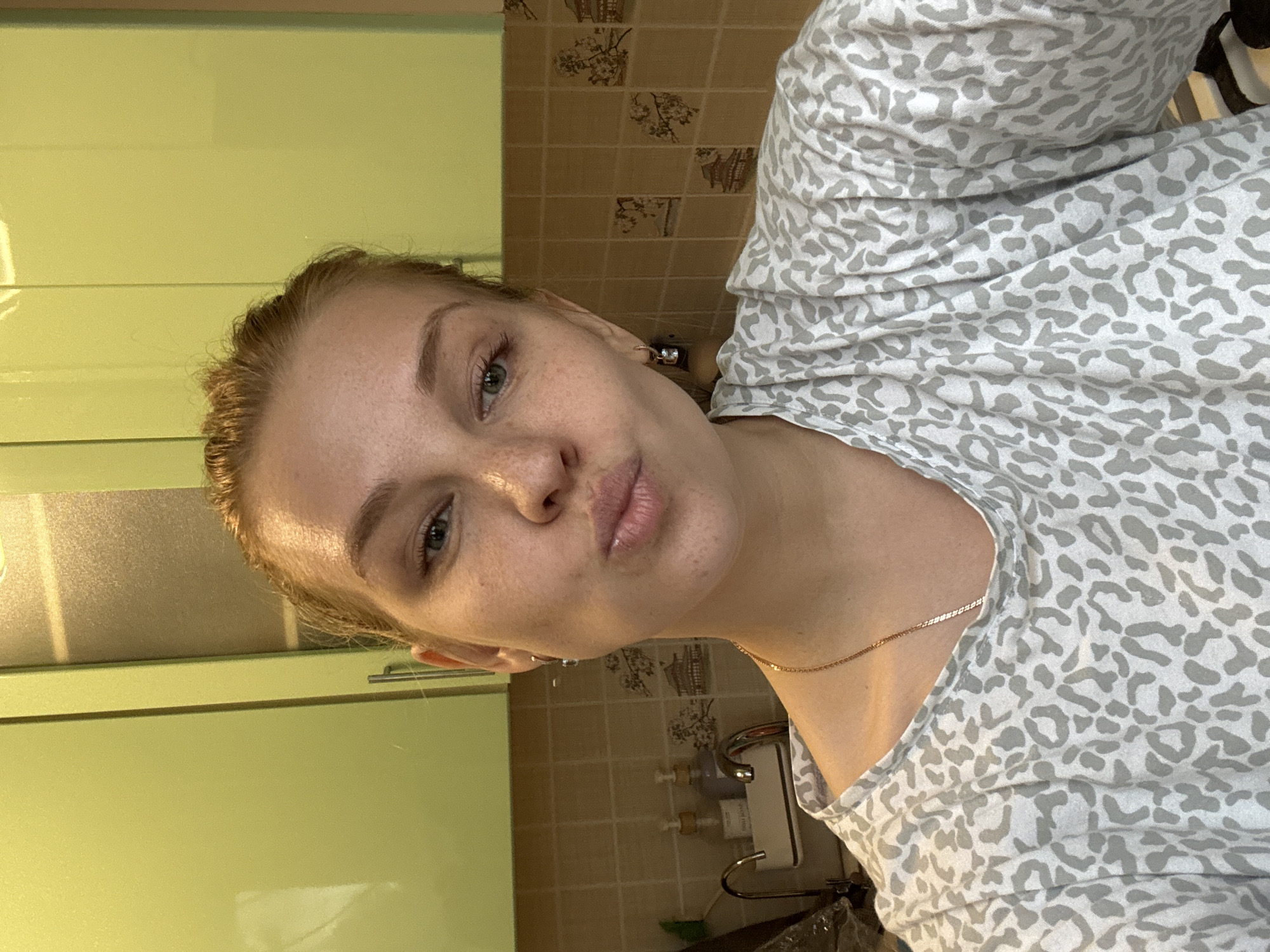 A woman is posing for a selfie indoors, likely in a kitchen or bathroom. The location could be Rusanovo, Russia, though this is not definitively discernible from the image alone. She is wearing a gray and white animal-print top and a delicate gold necklace. She has fair skin with freckles, light-colored hair pulled back from her face, and light eyes. She is puckering her lips slightly.
The background shows green kitchen cabinets and beige tiled walls with a floral pattern. A sink, faucet, and soap dispenser are visible near the lower right of the frame.