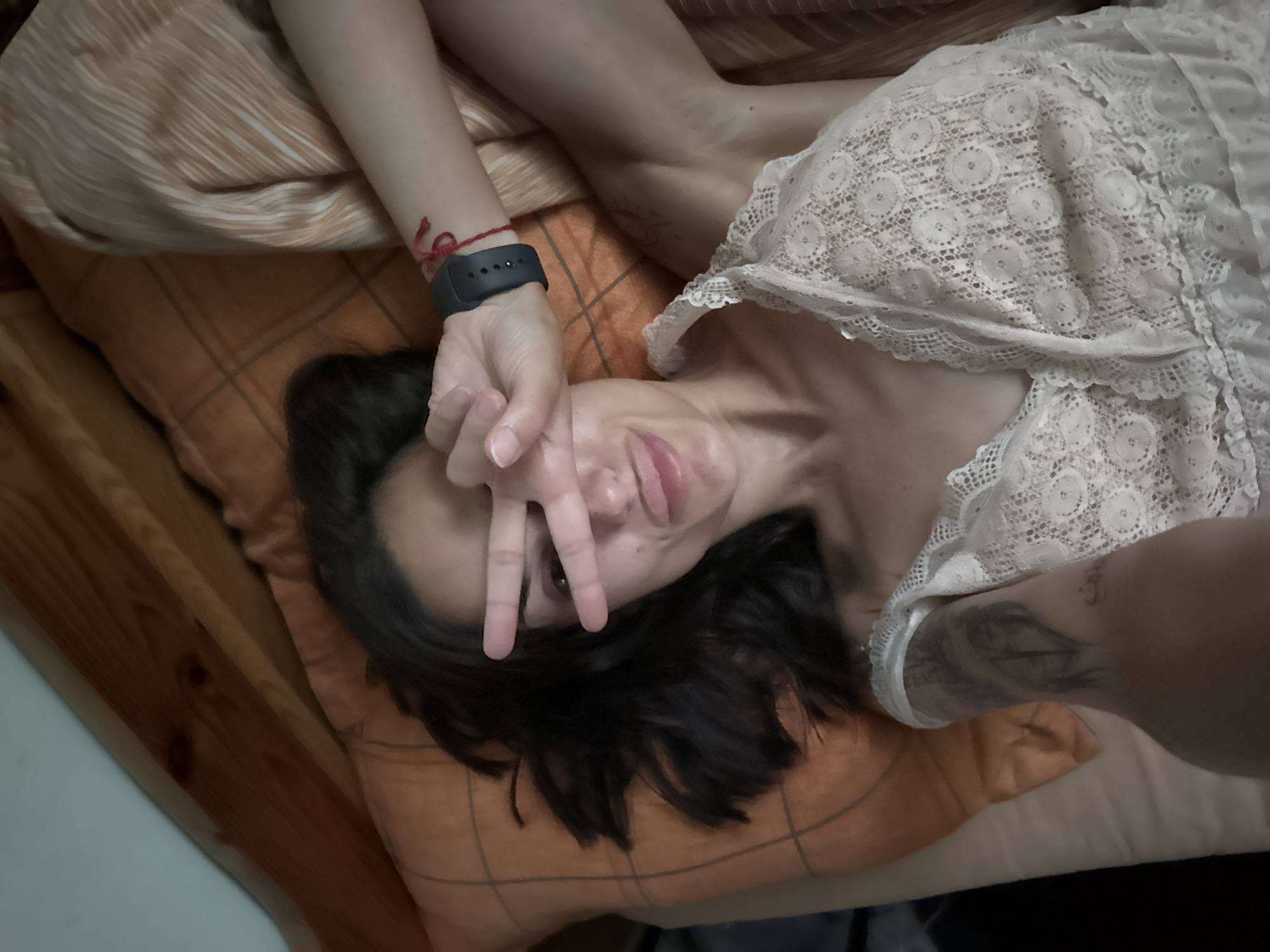 A woman is lying on a bed, with her head resting on an orange pillow. She is looking at the camera and holding up her hand to cover her eyes, making a peace sign. She is wearing a white lace top. The background consists of a wooden headboard and a patterned duvet. The scene is indoors, likely in a bedroom. The location is not specified in the image.