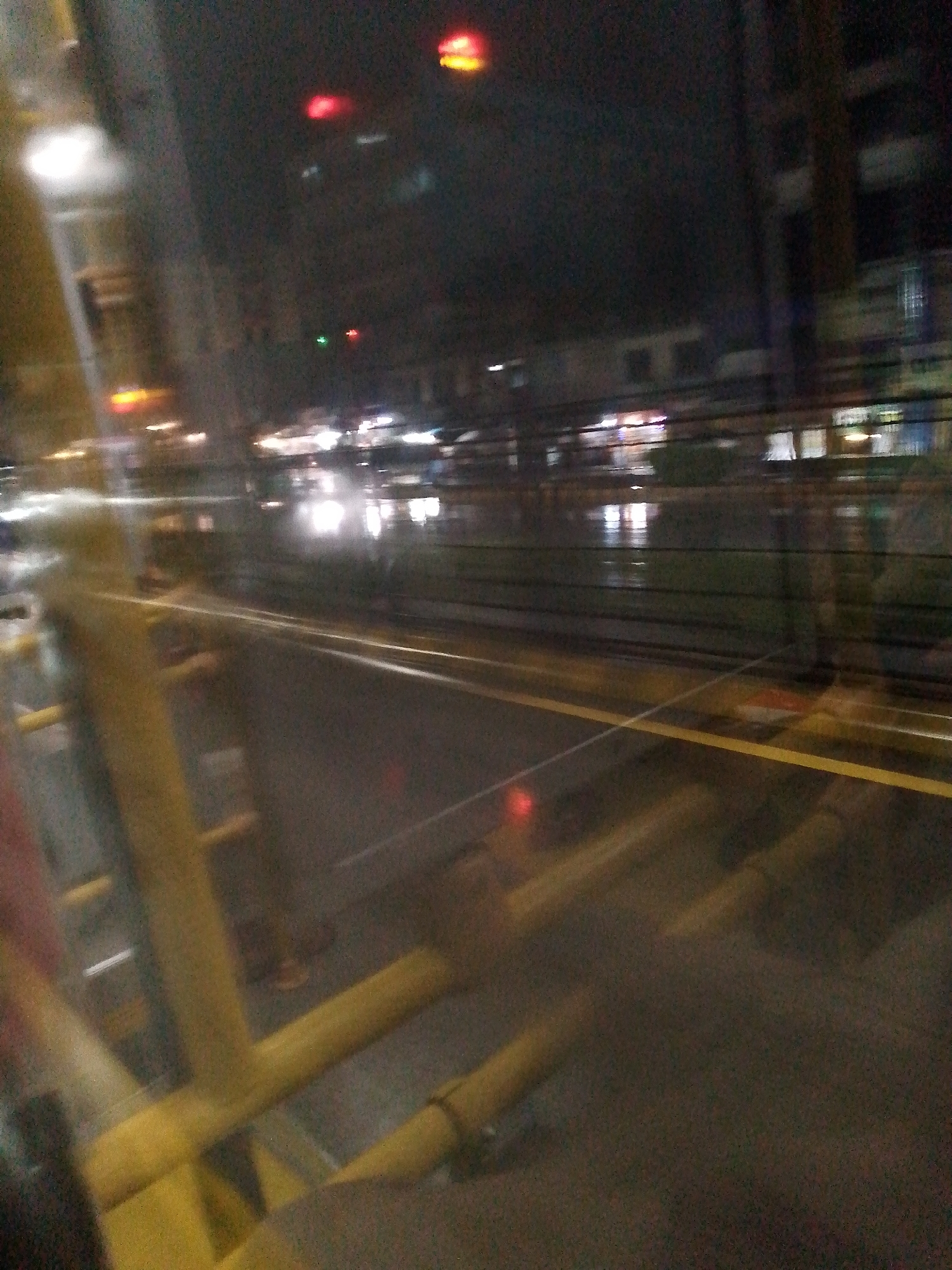 The scene depicts a street view at night, possibly taken from inside a vehicle or elevated position due to the presence of yellow metal structures in the foreground. The street is wet, reflecting the lights of buildings and street lamps. In the distance, there are buildings and indistinct shapes, possibly indicating commercial establishments, based on the spread-out lights. There are some blurred street lights, traffic lights, and other road objects. The street appears to be a wide thoroughfare with multiple lanes. The overall environment is dark with areas of illumination from artificial light.