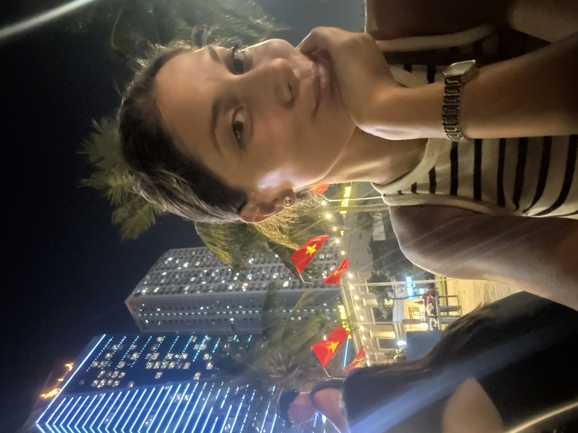 A rotated, low-angle, close-up portrait photograph of a woman with dark hair, fair skin, wearing a striped tank top and metal wristwatch. Her hand is near her face in the right-center of the image. She is looking towards the camera. In the background, illuminated buildings suggest a modern city environment. Red Vietnamese flags are visible. The city lights, flag color, and modern architecture suggest the location is Đà Nẵng, Vietnam. Palm trees are also present in the scene. Other people are partially visible in the lower portion of the photograph.