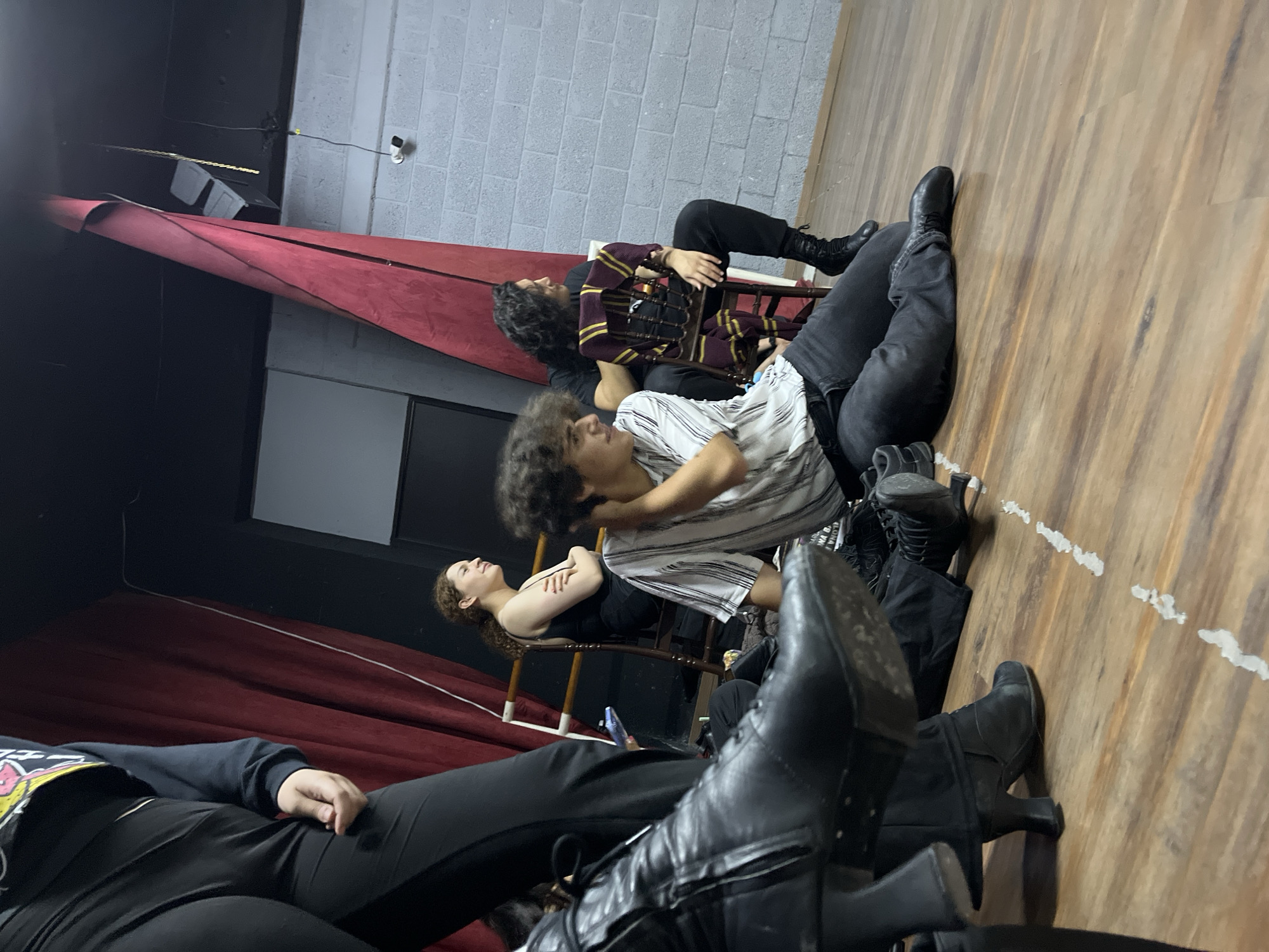 The image depicts a group of people resting on the floor and chairs in what appears to be a rehearsal or performance space. The flooring is wooden. A red curtain drapes in the background. The walls are a combination of brick and black material. One person wears a maroon and gold striped top. Several people wear black clothing. A woman reclines in a chair, eyes closed. Another person is dressed in a horizontally striped shirt. The location is unidentified.
