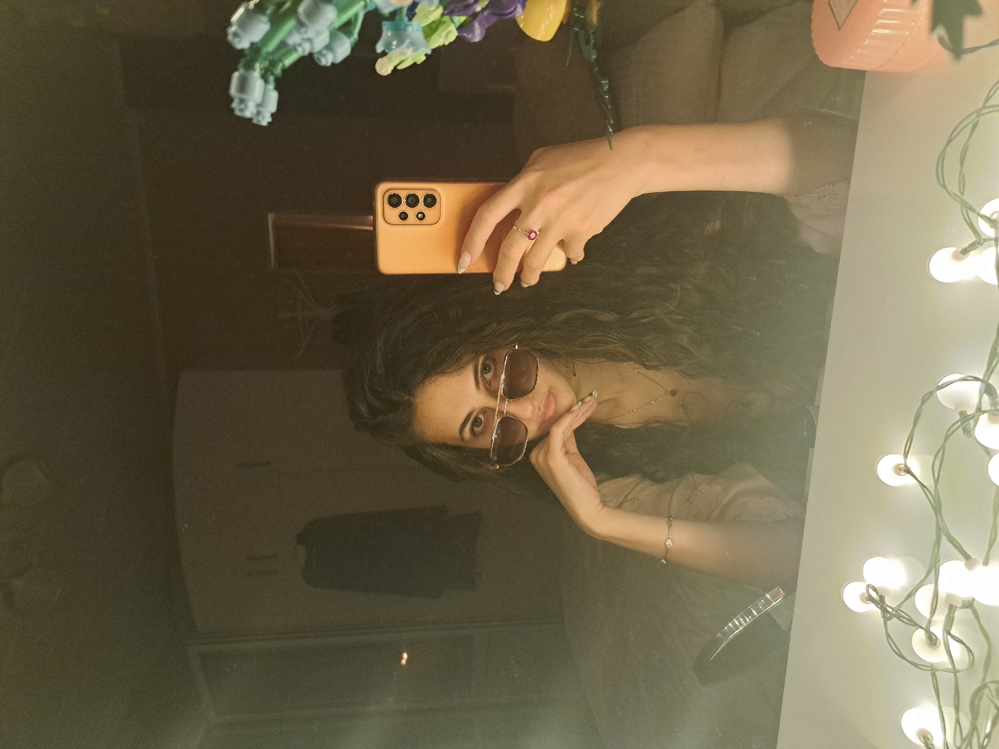 A mirror selfie captures a young woman posing indoors. The woman has long, dark, curly hair and is wearing sunglasses. She holds an orange-colored smartphone in her right hand to take the photo, while her left hand rests near her chin. A ring with a red gemstone is visible on her left ring finger, and a delicate bracelet adorns her right wrist. Above her reflected hand, artificial light blue and purple flowers along with a yellow decorative item are visible. To the right, a string of glowing white fairy lights is draped. The reflection in the mirror reveals the interior of a room, likely a bedroom or living space, with furnishings such as a light-colored sofa or bed. A pink cylindrical object is partially visible in the upper right. The woman is looking directly into the mirror, presenting a posed expression. No discernible text is visible within the image.