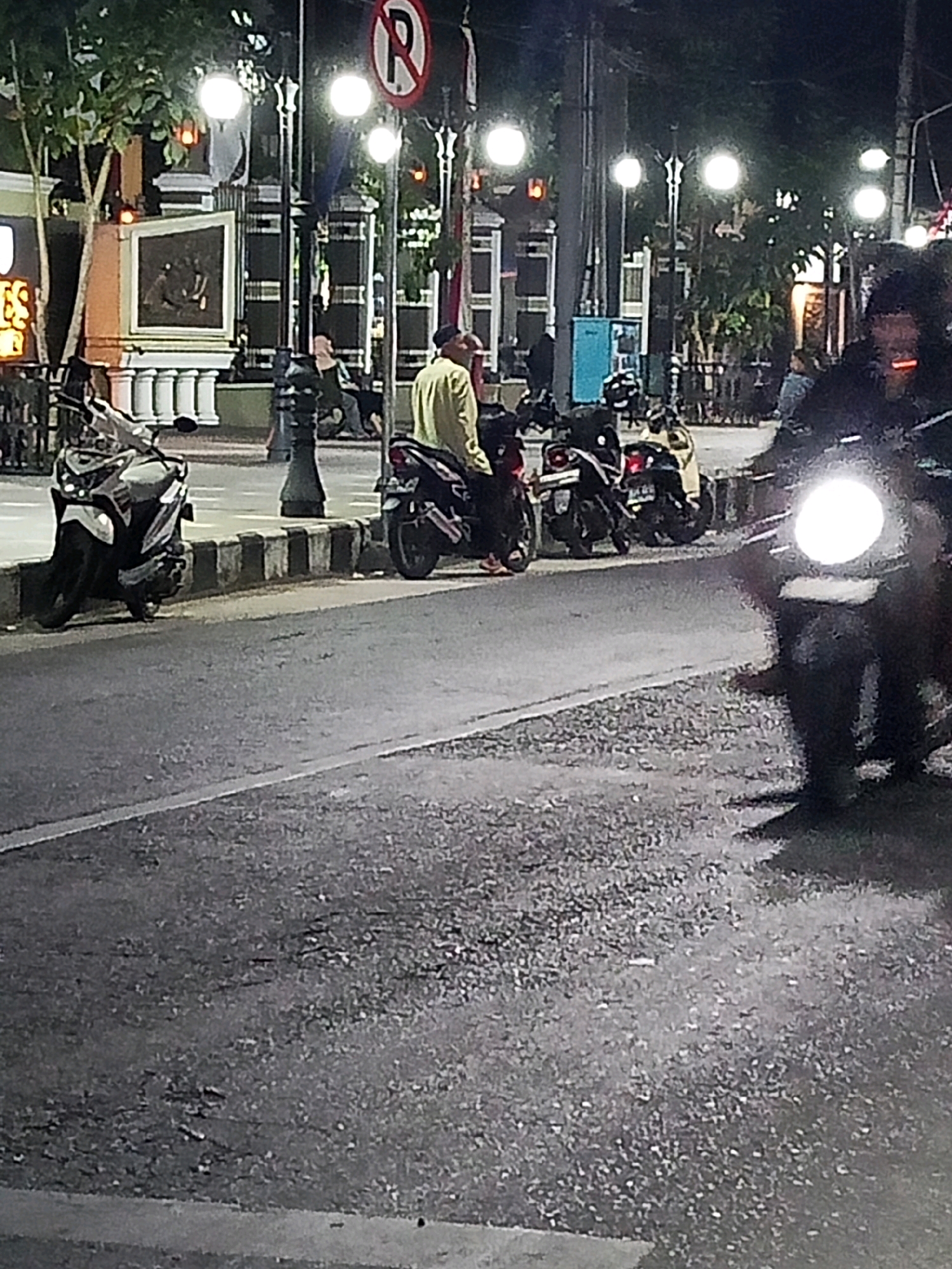 The image depicts a nighttime street scene in Wates, Indonesia. In the foreground, a dark-colored motorcycle with its headlight illuminated is the most prominent subject. Its front wheel and part of the rider are visible. Further back, parked along a curb, are several other motorcycles. One is a white and silver scooter on the left, with its kickstand down. To its right, a dark motorcycle is parked, and on it sits a person wearing a light-colored long-sleeved shirt and dark pants. Another dark motorcycle is parked next to that one. In the background, streetlights with bright bulbs illuminate the scene, casting a warm glow. Trees with lush foliage are visible behind the streetlights, and some architectural elements, including pillars and a framed artwork or plaque, are present on the left side of the image. A "no parking" sign is visible on a pole in the upper left quadrant of the image. The road surface appears to be asphalt, with a striped curb separating the road from the sidewalk area where the motorcycles are parked. The overall atmosphere is that of a quiet urban evening.