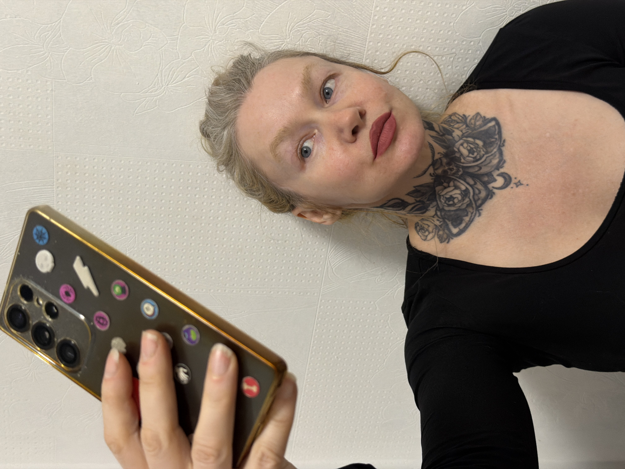 A Caucasian woman with graying blonde hair and blue eyes is holding a smartphone and taking a selfie. She is wearing a black top and has a floral tattoo on her neck. The smartphone has a gold-colored case and is decorated with various stickers. The background is a white wall with a textured floral pattern. No information suggests that the location is Saint Petersburg, Russia.