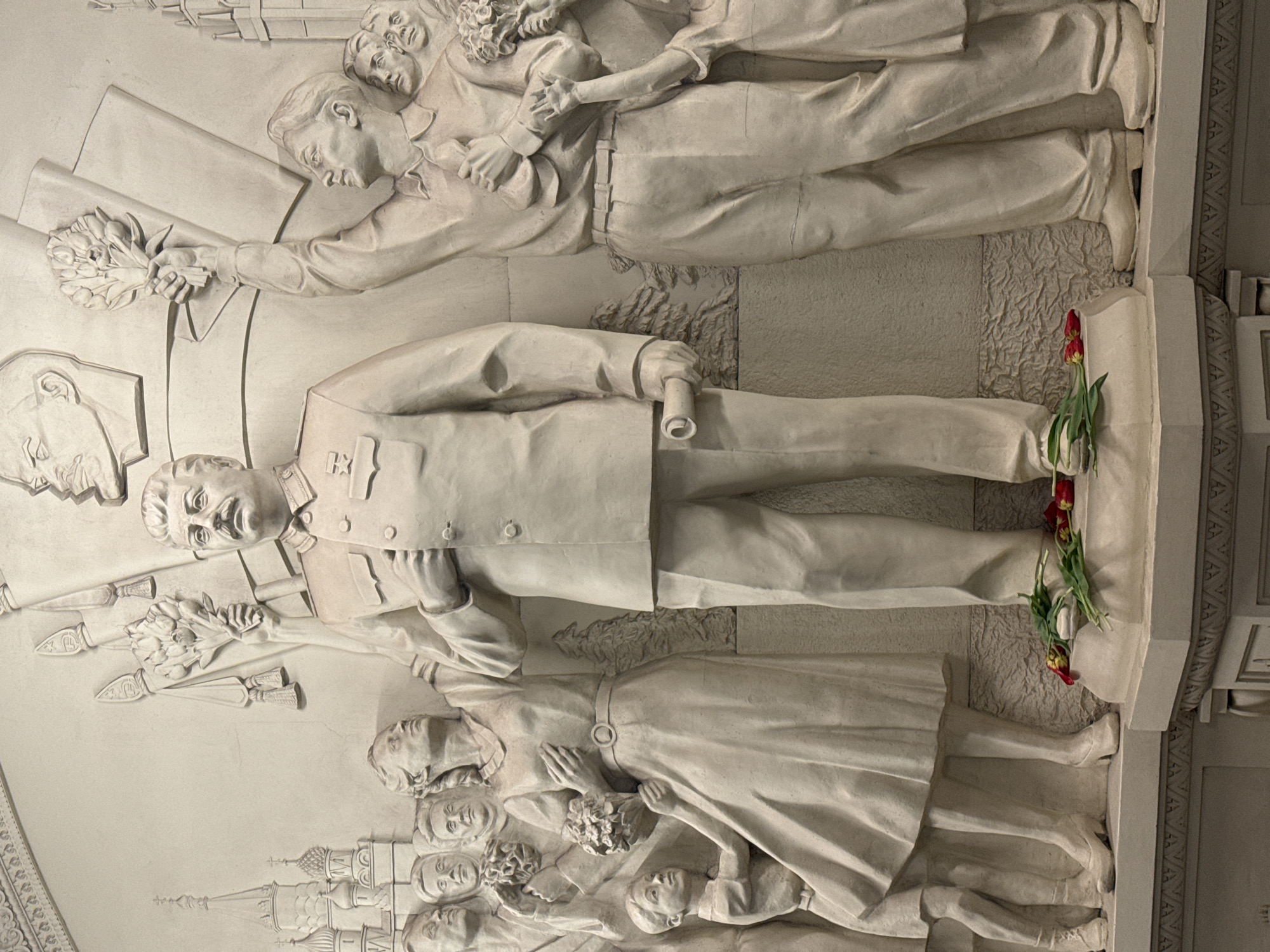 The image shows a white bas-relief sculpture on a wall. The central figure is a standing man in a suit, likely intended to be Joseph Stalin, judging by the facial features and uniform details. The sculpture includes other figures, including people carrying flowers and the depiction of Moscow's Kremlin. Red tulips have been placed at the foot of the central figure. This is likely located in a metro station in Moscow, Russia.