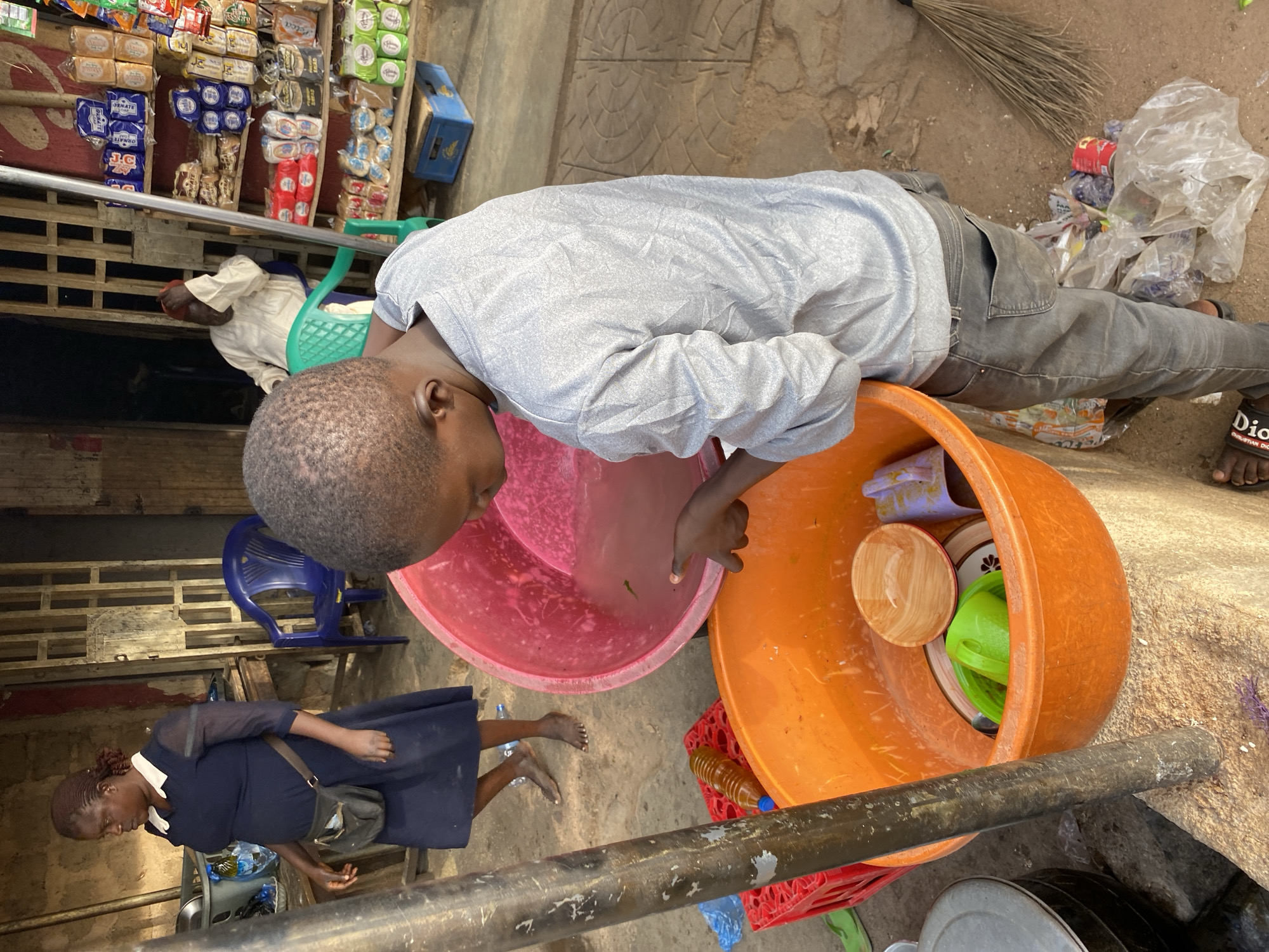 The image depicts an outdoor scene in Bukuru, Nigeria, likely a street market or vendor stall, captured during the daytime.
In the foreground, a young boy, appearing to be a child or pre-teen, is leaning forward over a large pink plastic tub filled with water, his hands submerged. He is wearing a light grey long-sleeved shirt and dark grey trousers. On his feet are black sandals, one of which clearly displays "CHRISTIAN DIO" (likely Christian Dior). Adjacent to the pink tub is a larger orange plastic tub containing various household items, including wooden and plastic bowls, and a green cup.
In the mid-ground to the left, a woman wearing a dark blue dress with a white collar and carrying a shoulder bag stands barefoot, looking downwards. Further behind her and partially obscured by a wooden shelving unit, another person in light-colored clothing is seated on a green plastic chair.
The background features a wooden market stall structure laden with various packaged goods, including loaves of bread, biscuits, and other products in blue and red packaging. Text such as "ORANGE," "DT," and "SWEET" is visible on some items. A blue plastic crate is also visible near the stall.
The ground is a mix of concrete and packed earth, showing some textured patterns. To the right of the boy, a pile of plastic bags containing various items and a natural-fiber broom are visible. A metal pipe or railing runs horizontally across the lower part of the frame. A plastic bottle with a yellowish liquid rests on the ground near the orange tub. The scene conveys an everyday moment of activity in an open-air setting.