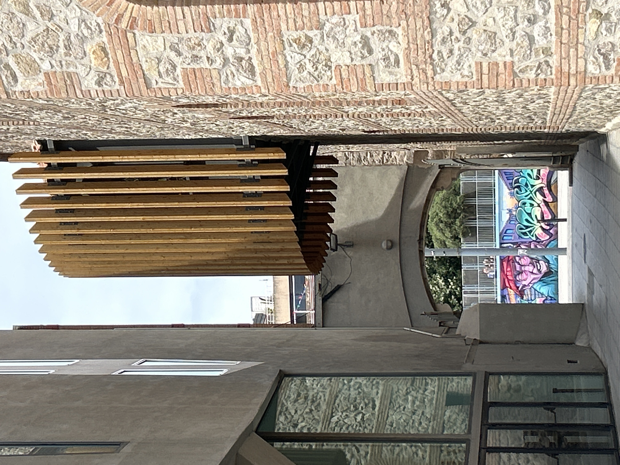 This is a medium-angle, eye-level, outdoor shot of an alleyway in Barcelona, Spain, on a cloudy day. On the right is a wall constructed of rough, light-colored stones and red brick. To the left of the stone wall is a modern architectural feature made of vertical wooden slats. The left side of the alley is bordered by a gray building with several rectangular windows, one of which reflects the stone wall and wooden slats. The alleyway continues into the distance where there is a large archway. Underneath the archway, a section of a wall features colorful graffiti depicting a cartoonish figure with a red bandana and a bright blue and green abstract design. A white railing is visible above the graffiti. A light pole stands in the foreground on the right side of the alley.
