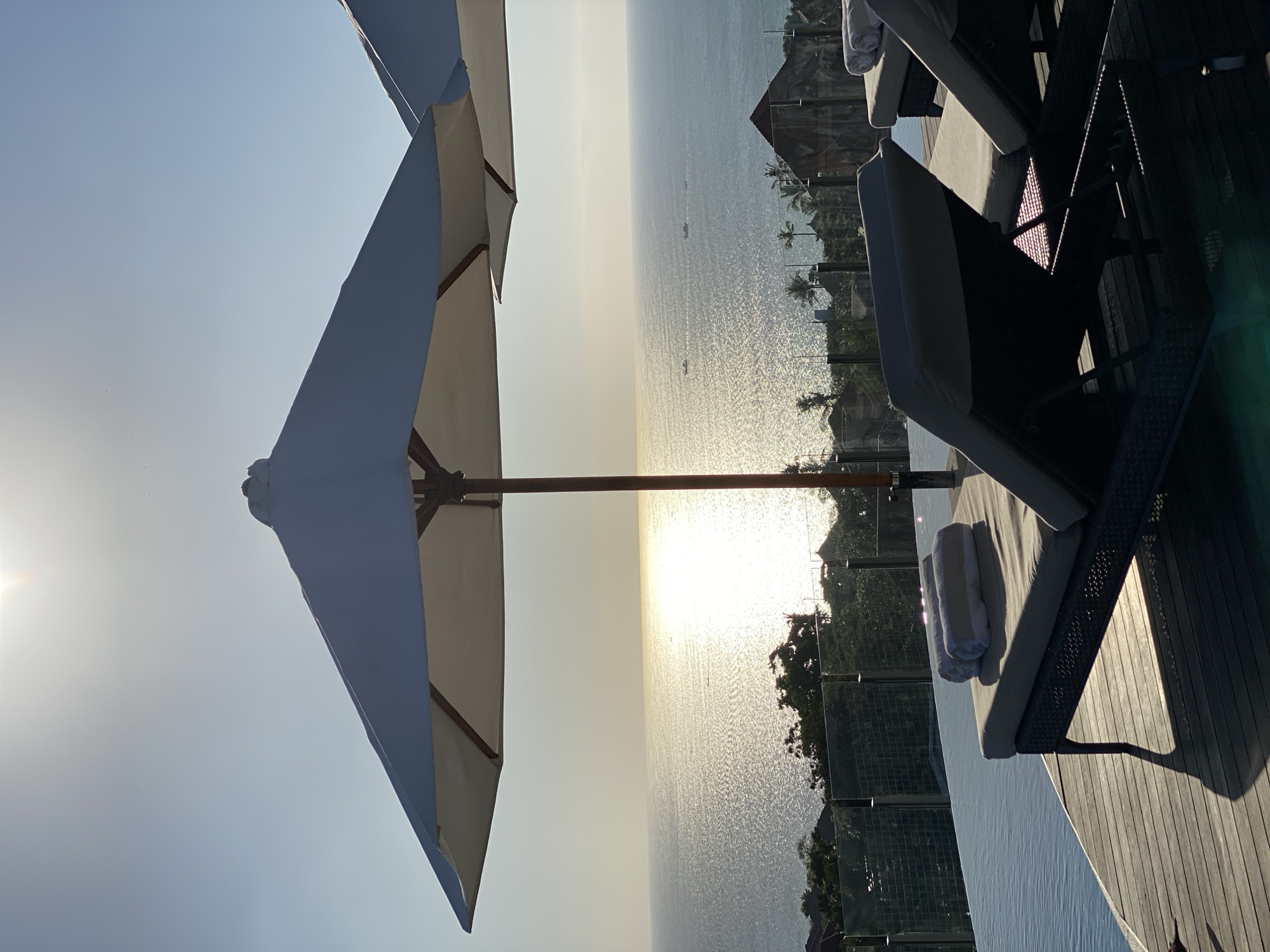 This image captures a serene poolside scene in Kuta Selatan, Indonesia, likely at a resort or villa. The foreground features two empty lounge chairs positioned on a wooden deck next to an infinity pool. Each chair has rolled towels placed on it. A large white umbrella provides shade over one of the chairs. In the background, the vast expanse of the ocean stretches to the horizon, reflecting the bright sunlight. A line of trees and some structures are visible on the coastline below, with a few small boats dotting the water. The sky is clear and pale blue, with the sun prominently positioned in the upper left quadrant. The overall atmosphere is tranquil and suggestive of a tropical vacation.