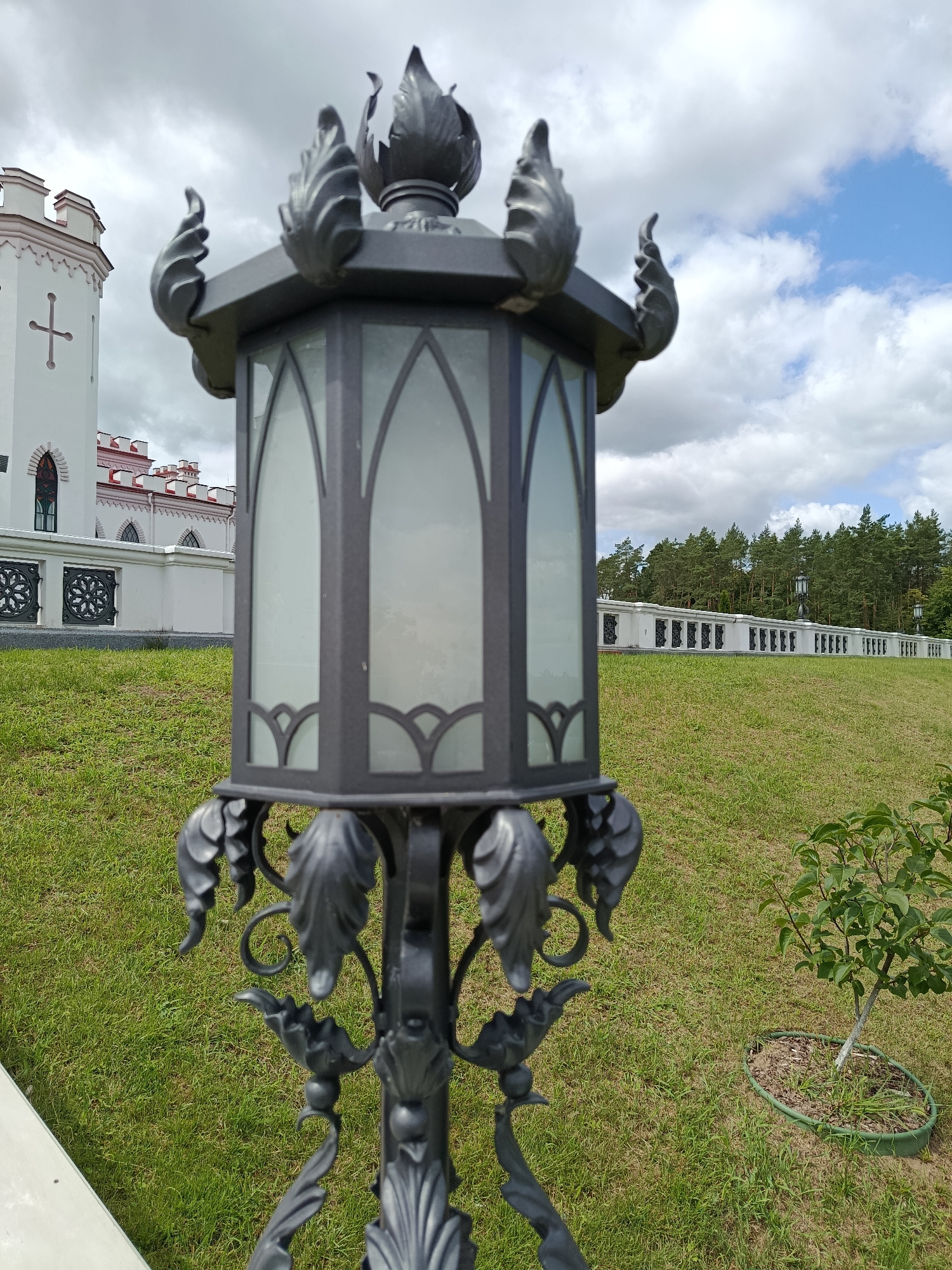 This is a medium shot, eye-level, outdoor photograph of a decorative lantern in the foreground, with the Kosava Palace (also known as Puslovsky Palace) in the background. The lantern is black and ornate, with glass panels and flame-like embellishments at the top. It is positioned on a grassy lawn. Behind the lantern, a white building with red trim and crenellations is visible. The building has arched windows and a cross emblem. A white fence with decorative black metal inserts runs horizontally across the middle ground. A line of trees forms the background, with a cloudy sky overhead. The overall scene depicts the grounds of the Kosava Palace in Kosava, Belarus. There are no people or animals in the image.