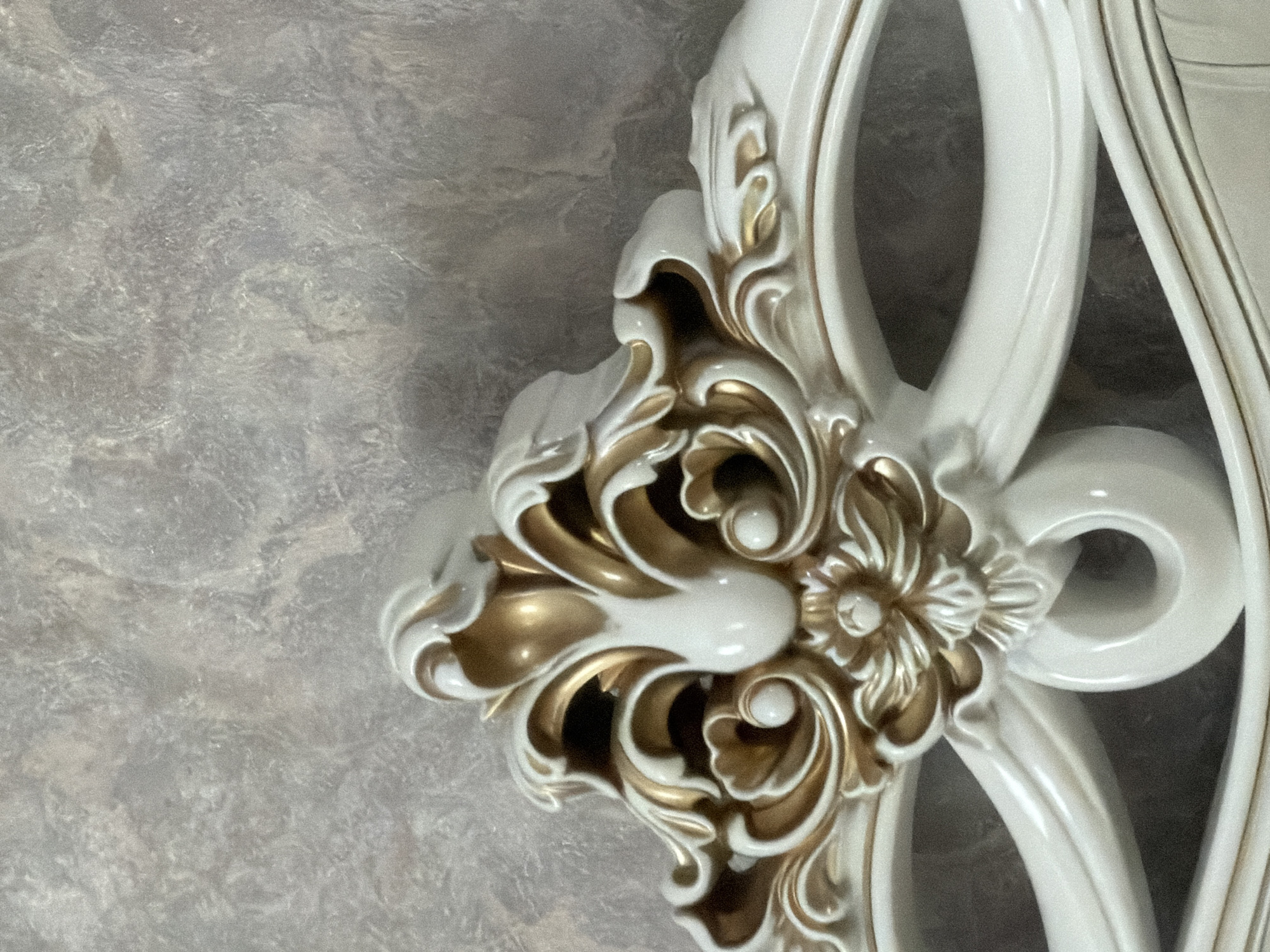 This is a close-up, eye-level, medium shot of an ornate decorative element, likely a furniture crest or architectural molding, rendered in white with gold accents. The material appears to be a resin or carved wood with a glossy finish. The design is elaborate, featuring acanthus leaf motifs, scrolled embellishments, and a central floral element. The background is a textured wall, possibly wallpaper or plaster, in muted shades of grey and beige. The lighting is soft, casting subtle shadows that highlight the depth and detail of the decorative piece. The image is devoid of any people, animals, or signs of activity. The precise location in Karshyga-Aul, Russia cannot be determined from the visual information provided.