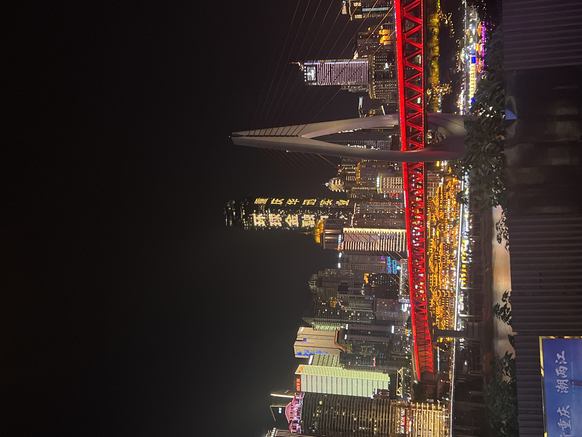 This is a night-time, medium-distance shot of a city skyline, likely Chongqing, China, given the presence of the landmark bridge and illuminated buildings. The scene is dominated by towering skyscrapers, many of which are lit up with various colors and patterns. The most prominent structure in the foreground is a modern cable-stayed bridge with a distinctive V-shaped pylon, painted red and illuminated with red lights. This bridge spans a body of water, reflecting the lights of the city. On the far side of the water, there are more buildings, some appearing to be older, traditional structures with illuminated roofs, clustered together. A tall skyscraper on the left side of the image has vertical text in yellow, likely a company or building name. The sky is dark and clear, providing a stark contrast to the vibrant city lights. In the lower foreground, a dark metal fence partially obscures a sign with Chinese characters and blue and white text. No people are visible in the image.