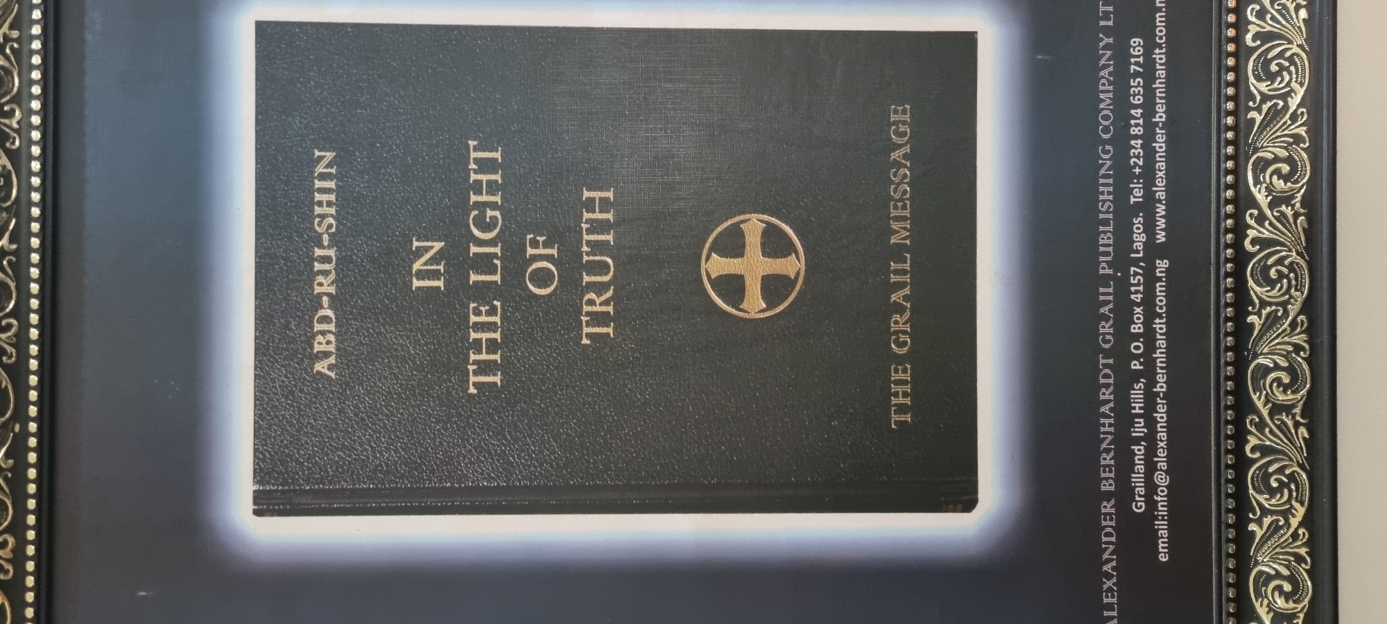 The image shows a close-up of the cover of a book titled "IN THE LIGHT OF TRUTH" by ABD-RU-SHIN. The book cover is dark, with gold lettering and a gold symbol resembling a cross within a circle. Below the book title, the text "THE GRAIL MESSAGE" is also visible in gold lettering. At the bottom of the image, there is contact information for Alexander Bernhardt Grail Publishing Company Ltd., including an address at Grailland, Iju Hills, P. O. Box 4157, Lagos, Nigeria. There are no people or actions depicted in the image. The image is framed by an ornate border. The city of Ijaiye, Nigeria, is not directly visible or mentioned in the image.