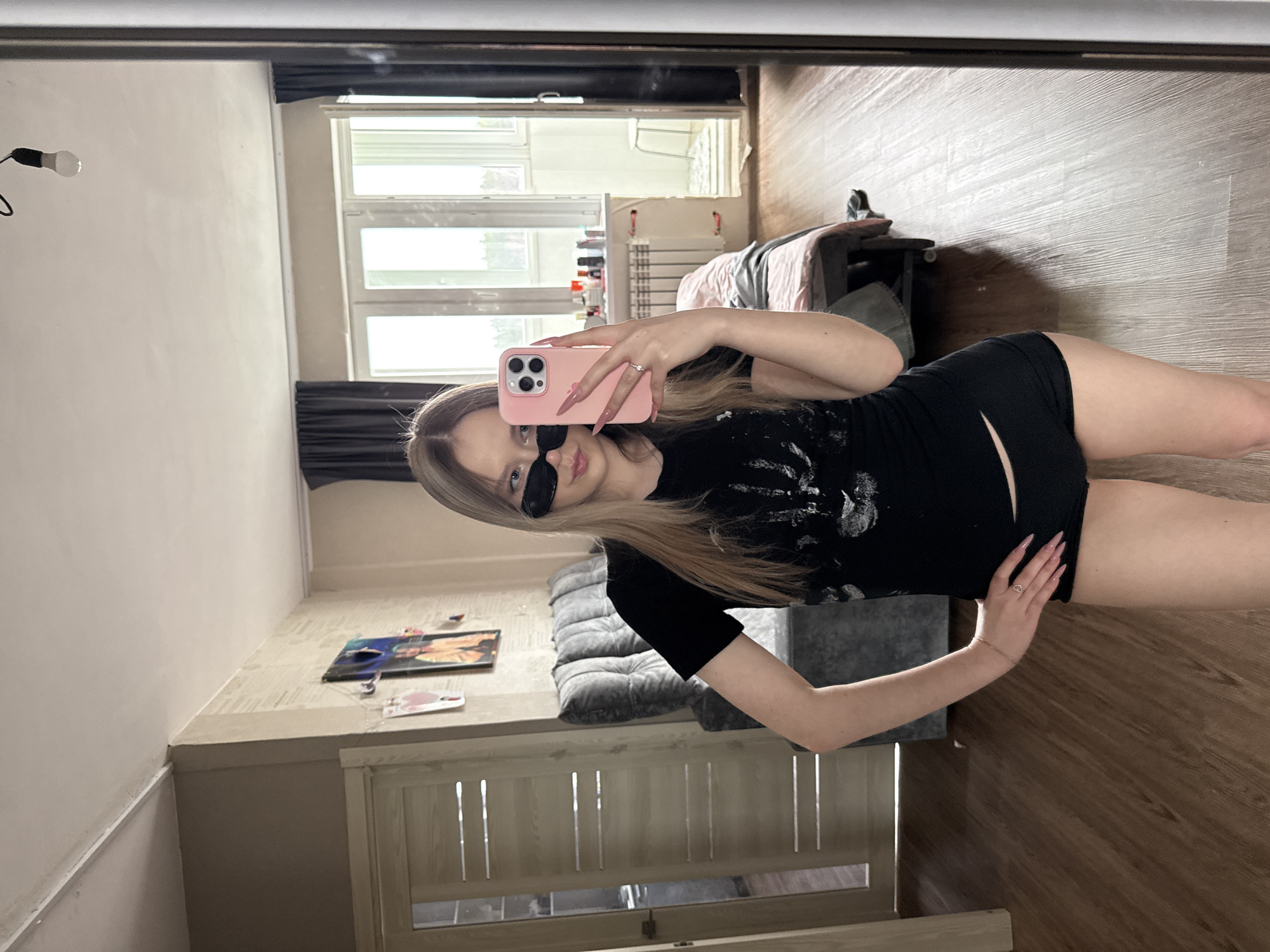A young woman with long blonde hair, wearing sunglasses and a black t-shirt with black shorts, is taking a selfie in front of a mirror. She is holding a pink phone in her left hand. The background includes a bedroom with a large window, a bed, and a couch. The location may be in Chaikovsky, Russia, based on the prompt. The room appears to be in a residential building.