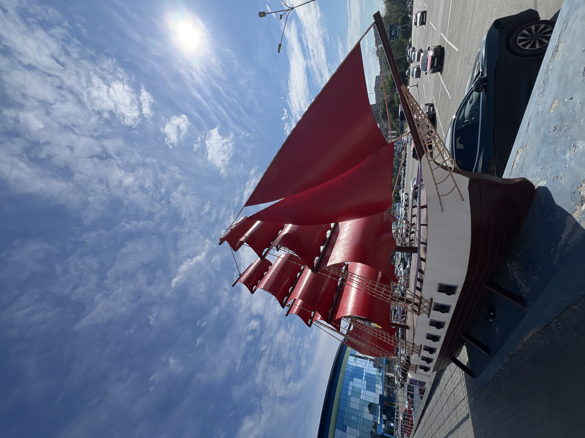 A large, decorative model of a sailing ship with bright red sails is positioned outdoors in a parking lot, facing a modern glass-fronted building. The ship model is dark brown and white, with multiple rows of sails. The sky above is blue with scattered white clouds and the sun is visible, casting a lens flare. Cars are parked in the lot behind the ship, and in the distance, there are trees and buildings. The ship appears to be an attraction or landmark in Izhevsk, Russia, situated in front of what looks like a commercial or entertainment venue.