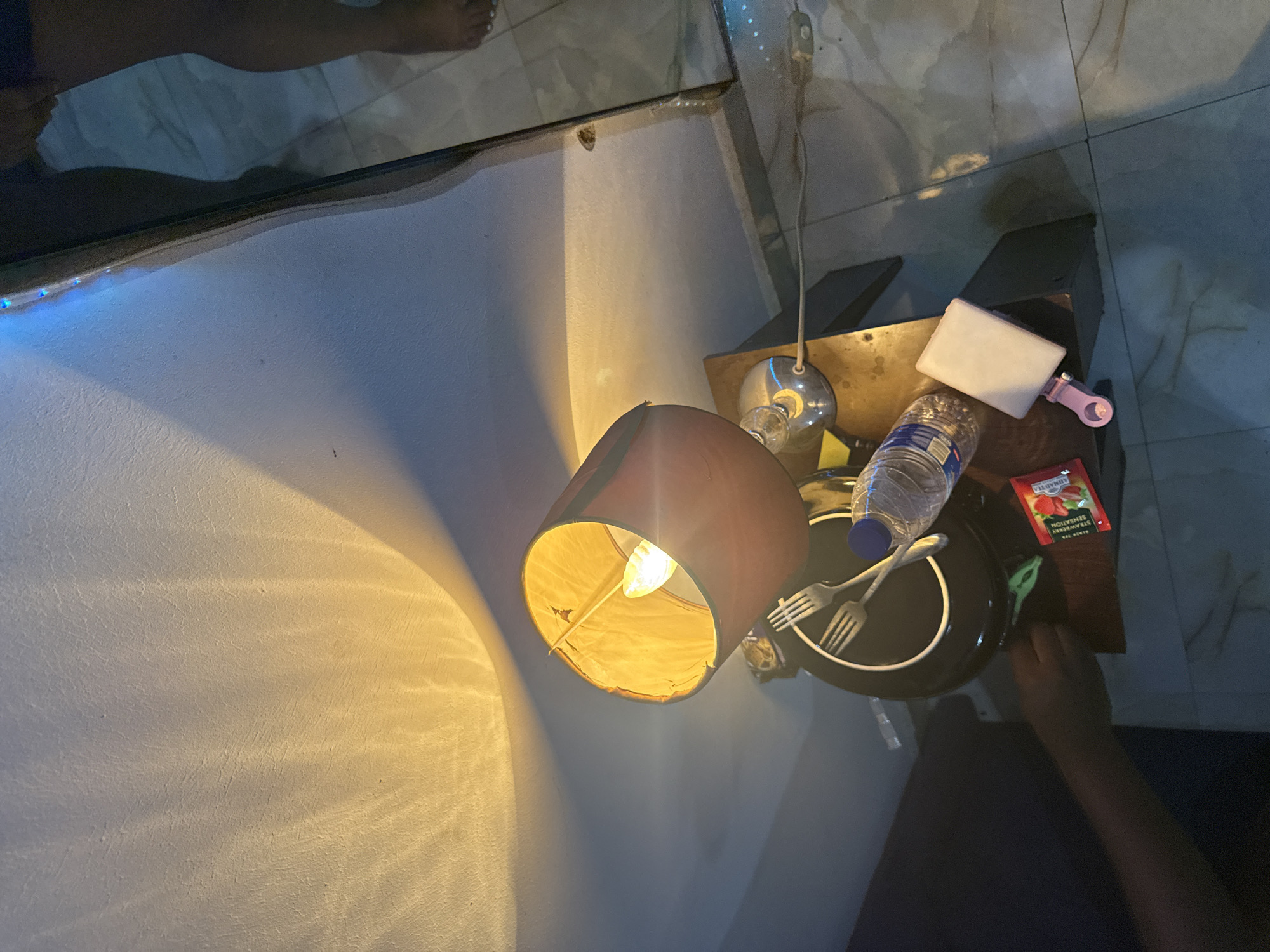 The image shows a dimly lit room, possibly in Abuja, Nigeria, based on the presence of a brand of tea called "Strawberry Sensation" which is commonly sold in Nigeria. A lamp with a brown shade casts a warm light onto a white wall. On a small wooden table, there's a black plate with two forks, a partially visible water bottle, a small white container with a pink clip, and a packet of "Strawberry Sensation" tea. A person's arm is visible on the right side of the frame. The room appears to be tiled, and a partial view of feet is visible near what might be a mirror.