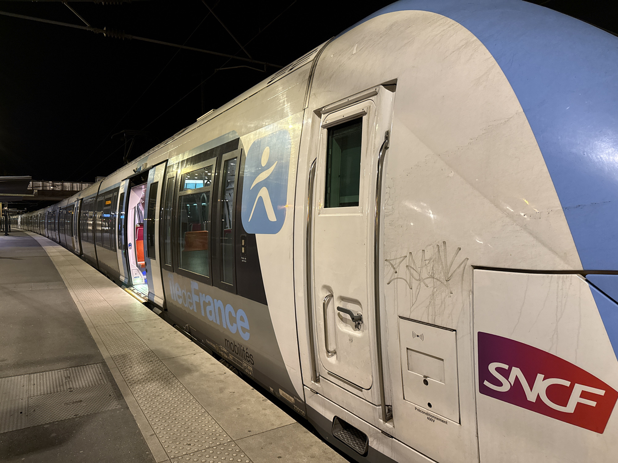 A nighttime view shows a white and blue SNCF train at a platform in Saint-Brice-sous-Forêt, France. The train's door is open, revealing a red interior. The side of the train has graffiti and logos for Île-de-France Mobilités and SNCF. The platform is visible in the foreground. Overhead electrical lines are visible above the train.