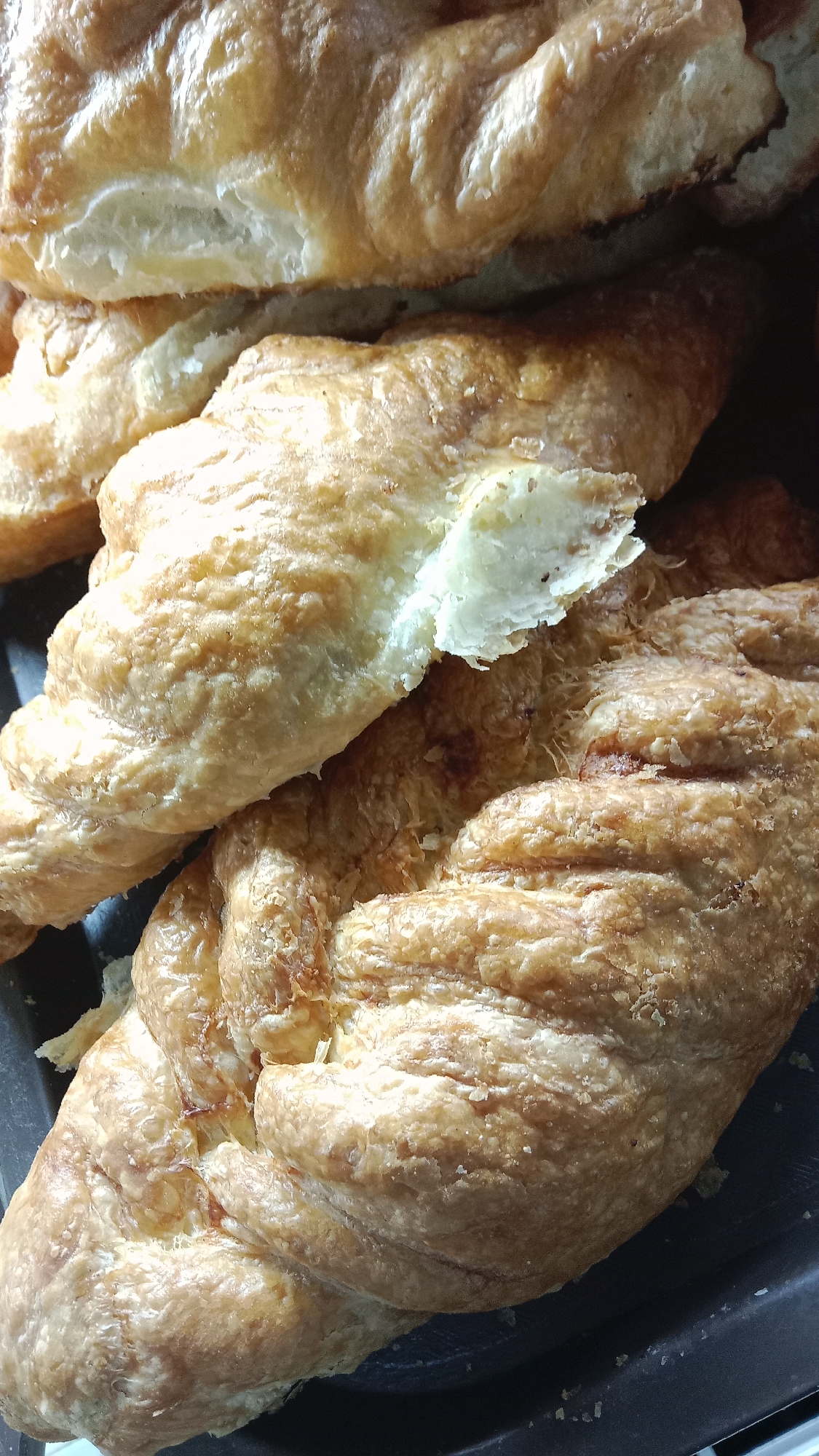A close-up, overhead shot shows a dark tray filled with golden-brown croissants. The croissants are stacked on top of each other, with the flaky pastry clearly visible. Some croissants are whole, while others appear to be broken or have pieces missing, revealing the airy interior. The lighting highlights the texture and sheen of the baked goods, suggesting they are fresh. The image does not contain any visual information that would allow for the identification of a specific location in Petropavl, Kazakhstan.