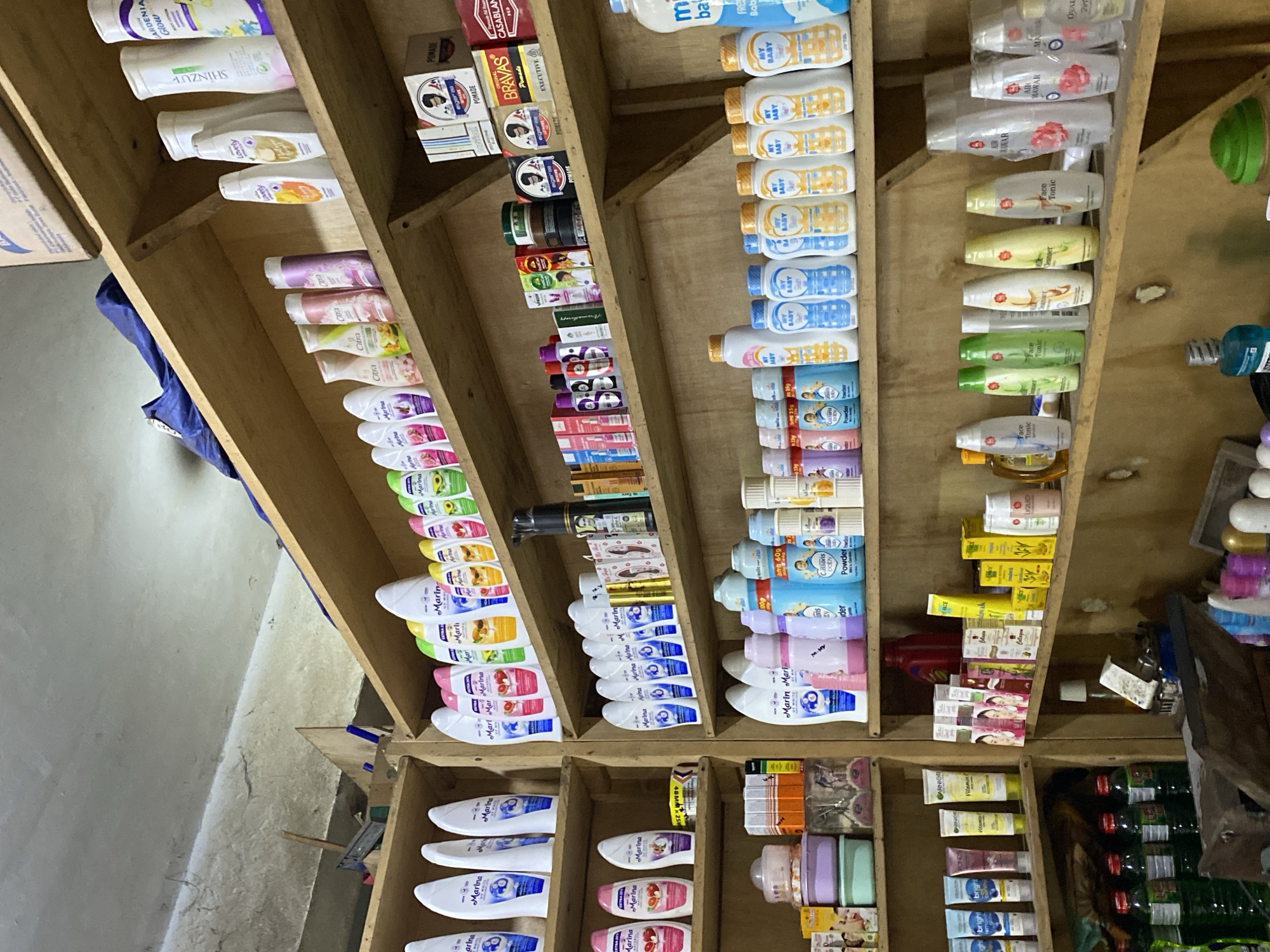 The image depicts the interior of a small shop, likely a convenience store or pharmacy, in Ta', Indonesia. The scene is dominated by shelves filled with a variety of personal care products, including lotions, powders, soaps, and cleansers. The products are predominantly from Indonesian brands, as suggested by the packaging and language visible on some items.
On the top shelf, there are several rows of lotions and body washes, with brands like Marina and Citra being prominent. Further to the right, there are smaller boxes of what appear to be hair pomade or styling products.
The middle shelves are also heavily stocked with personal care items. On the left side, there are rows of lotion bottles, and on the right, more lotions and body washes, along with some boxed products and canisters.
The bottom shelves show a mix of items. On the left, there are plastic containers and what appear to be food items or supplements in boxes. Moving to the right, there are more rows of baby powders and lotions, followed by bottles of liquids, potentially hand sanitizers or disinfectants.
The shelves themselves are made of light-colored wood, and the overall environment appears somewhat cluttered and utilitarian, typical of a small retail space. The lighting is adequate, illuminating the products clearly. No people are visible in the image. The context of Ta', Indonesia, suggests a local market setting.