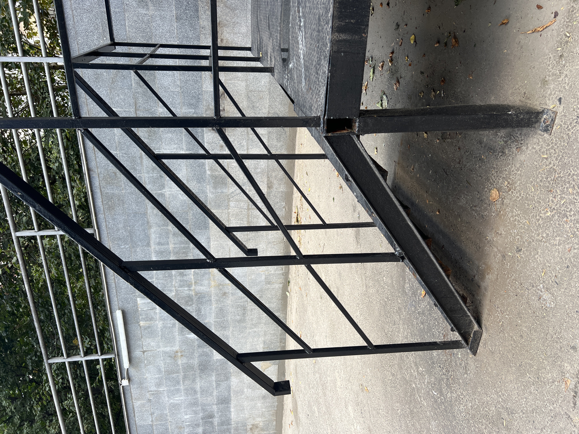 The image shows a close-up of a black metal staircase outdoors. The staircase has a textured metal platform and a handrail made of black metal bars. Behind the staircase, there is a grey brick wall and some greenery, likely trees. The ground in front of the stairs is paved with concrete and scattered with fallen leaves. The scene appears to be in a public outdoor space. There are no people or animals visible in the image. The lighting suggests it is daytime.