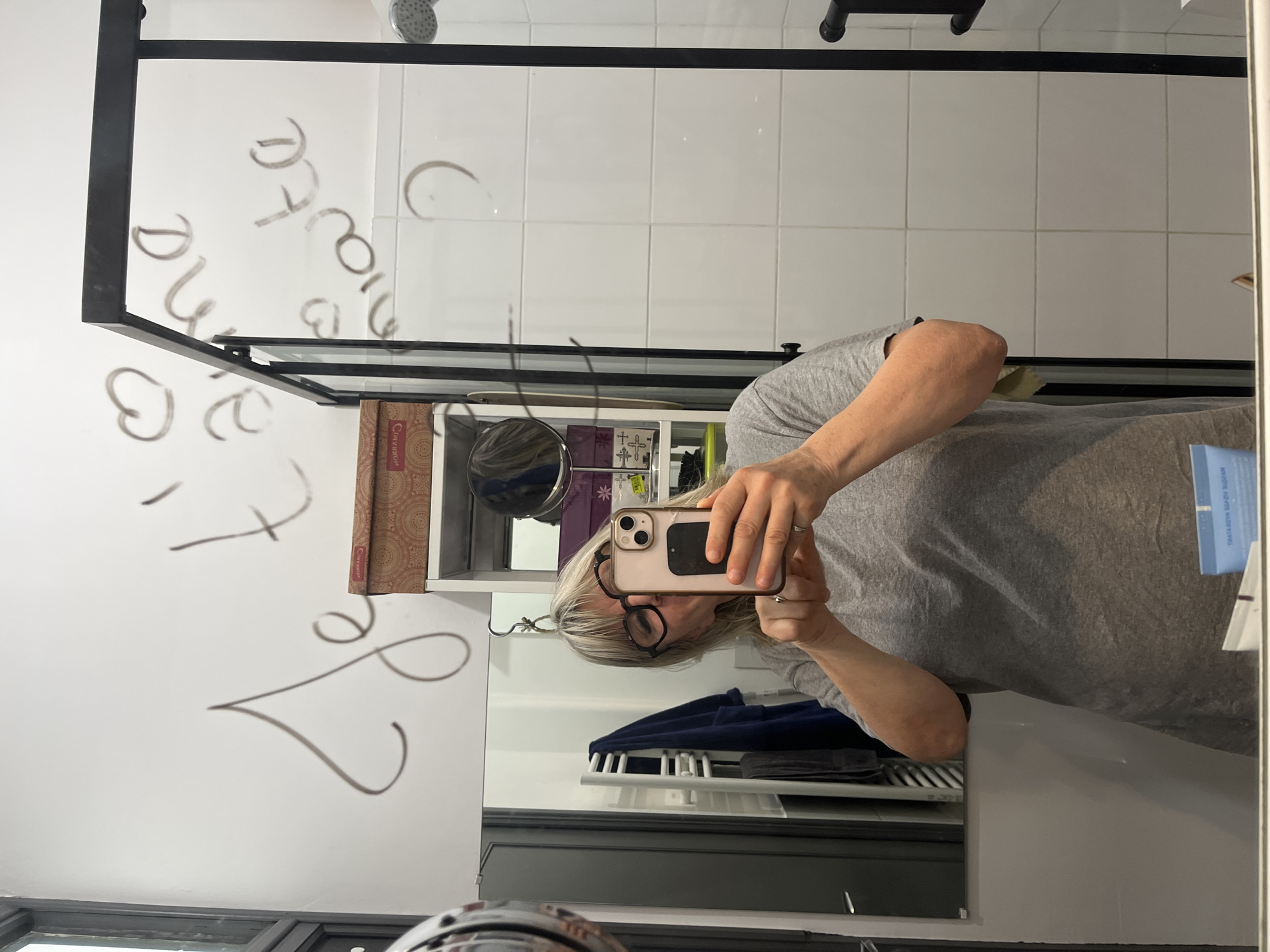 A person with blonde hair and glasses is taking a selfie with an iPhone in a bathroom mirror. The bathroom features white tiled walls, a shower head, and a black metal frame around a glass shower enclosure. The glass has writing on it. A shelf unit is visible in the reflection, containing various items. A towel rack with blue towels is also reflected in the mirror. The image was taken indoors. The location in the city of Montélimar, France, is not discernible from the image.
