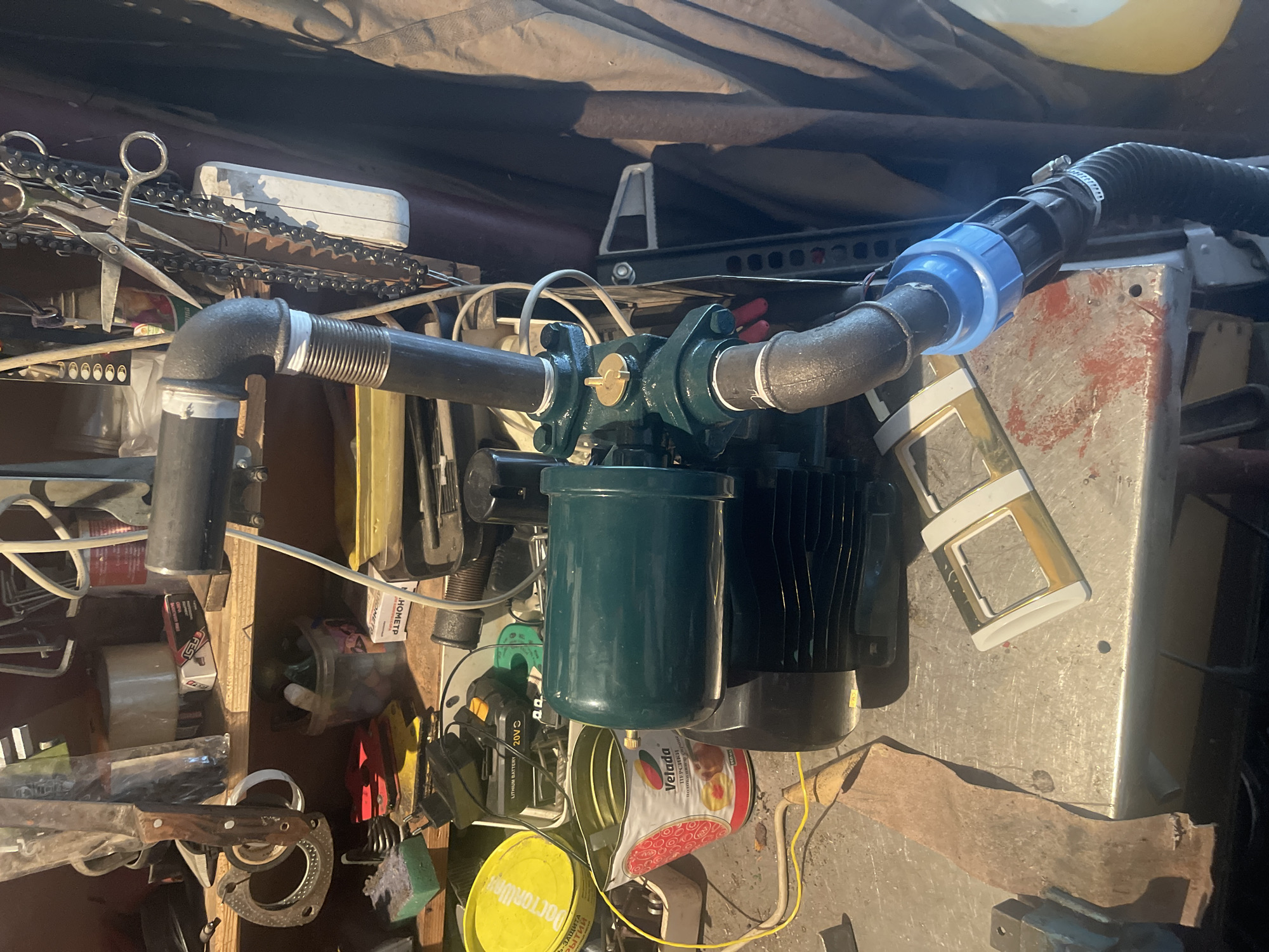 In the city of Sibirskiy, Russia, an image captures a cluttered workbench area, likely a garage or workshop, with various tools and equipment. A green submersible pump with black casing and metallic pipes is prominently displayed. The pump is connected to a flexible black hose secured with a white clamp and a blue fitting. To the left of the pump, a red and white can with a "Velada" label and illustrations of food items sits on the workbench. Behind the pump, various items are visible on shelves, including rolls of tape, scissors, a knife with a wooden handle, and other assorted tools. A yellow circular container with text is also present. The overall environment appears unkempt, with tools and supplies scattered around.
