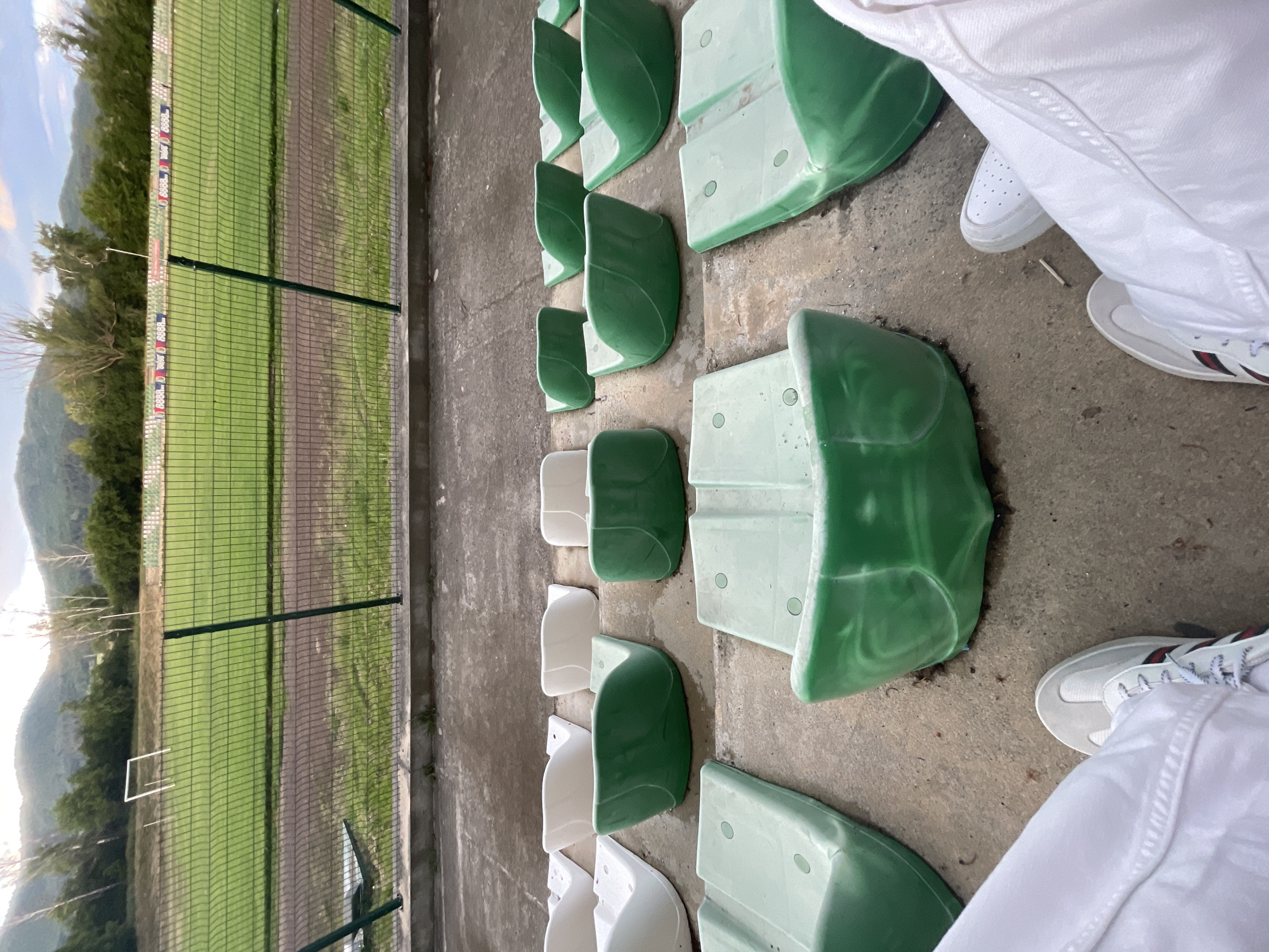 The image captures a scene from the stands of a sports ground in Ihtiman, Bulgaria. In the foreground, the lower body of a person is visible, wearing white trousers and white athletic sneakers, with the feet resting on the concrete ground.
The primary subjects are the rows of stadium seats, which are a mix of light green and white plastic, arranged alternately. These seats are mounted on a concrete surface, which forms the viewing area. A sturdy green mesh fence separates the spectator area from the playing field beyond.
In the mid-ground, past the fence, lies a green sports field or pitch. A distinct brown dirt path runs alongside the field, possibly a running track. In the far distance on the field, a small, white rectangular goal frame can be seen. Along the top of the fence, there are advertising banners featuring a repeated pattern of a red, white, and blue logo. While precise text is difficult to discern, a repeating numerical sequence resembling "8888" is part of the emblem on these banners.
The background features abundant green trees and undulating mountains under a bright blue sky with scattered clouds, suggesting a clear day. The overall lighting indicates daytime, possibly mid-morning or late afternoon, given the soft shadows. No specific activity or event like a match is actively taking place; the scene portrays the setting itself. The perspective is from a low angle within the stands, looking out towards the field and the distant landscape.