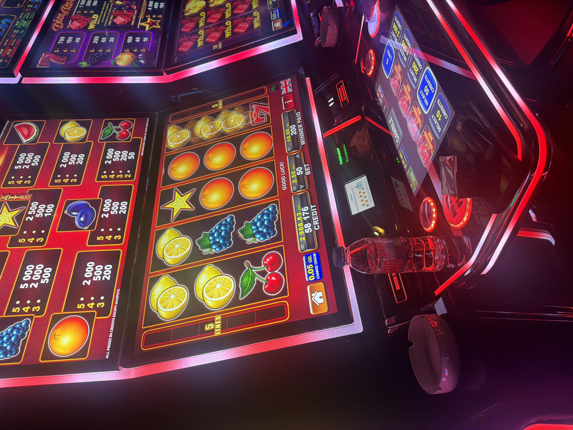The image captures a vibrant, illuminated scene within what appears to be a casino or gaming establishment, featuring multiple slot machines. The perspective is slightly elevated, looking down diagonally onto the machines and surrounding area.
The primary subjects are two active slot machines, prominently displaying fruit-themed symbols such as lemons, oranges, grapes, cherries, watermelons, and plums, along with classic slot symbols like "7"s, stars, and "WILD" icons. Payout tables are visible on the left machine, detailing credit amounts for various symbol combinations (e.g., 5000 for five watermelons, 2000 for five lemons). On the main visible machine, a digital display shows "2908.83 GEL" and "58 176 CREDIT," indicating a balance. Other game information includes "GOOD LUCK!," a "2.50 GEL" bet, and "10.00 GEL 200 WINNER PAID." The text "0.05 GEL CHANGE DENOM" is also visible. Buttons for "GAMBLE," "AUTO PLAY," and "SPEED" are seen on the right machine's interface, alongside options to "SELECT" different credit amounts (10, 50, 200, 1000, 2000). A "MALFUNCTION VOIDS ALL PAYS AND PLAYS" message is present on one screen.
Activity implies someone is currently playing or has just finished playing, as indicated by the active screens and displayed winnings. A plastic water bottle and a white ashtray are placed on the surface between the machines, suggesting a player is present and potentially taking a break.
The setting appears to be indoors, likely in the evening or at night, given the reliance on artificial lighting, characterized by bright red and white neon accents from the machines and their housing. The overall mood is one of focused entertainment. Visible text includes "Games Technology," "ALL PRIZES IN CREDITS EXCEPT JACKPOTS," "5 LINES," and the word "ИВЕРИА" (IVERIA), which could be a casino brand. A British flag is also displayed on one of the interfaces. The currency "GEL" indicates the location is likely Georgia.