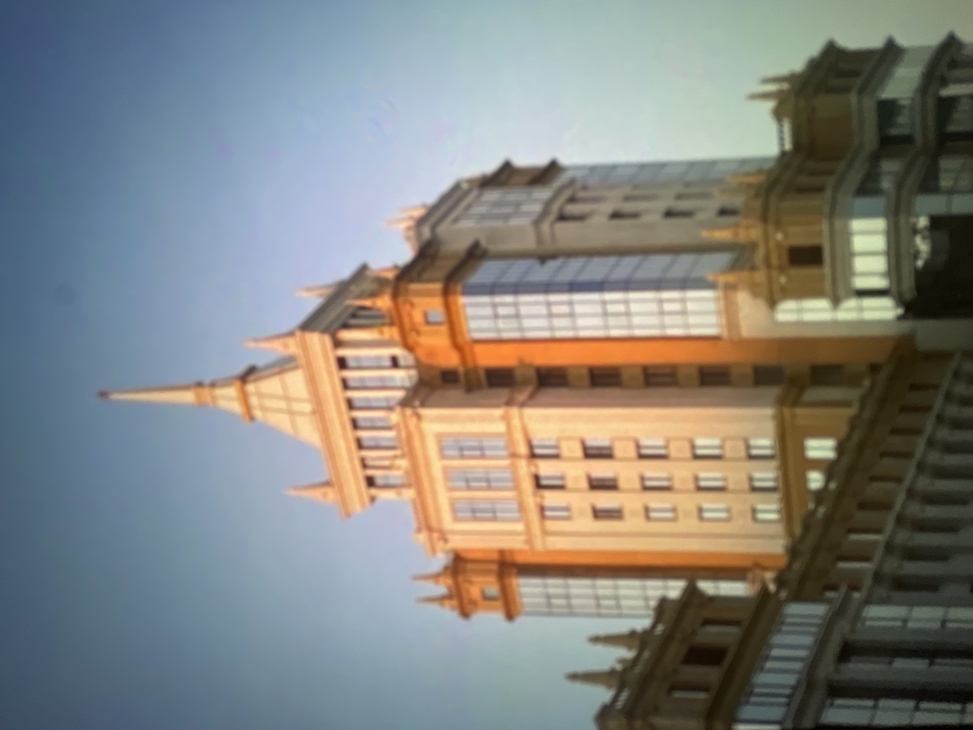 An eye-level, medium shot captures the upper section of a tall, ornate building against a clear blue sky. The building, rendered in warm tones of orange and beige, features a central spire and several smaller turrets. The facade is a mix of solid panels and windows, with a prominent section of blue-tinted glass. The architecture appears to be in a classical or Stalinist style. In the foreground, the tops of other buildings are visible, also displaying classical architectural elements. The image is slightly blurred, giving it a soft, almost dreamlike quality. The lighting suggests it is either early morning or late afternoon, with the sun casting a warm glow on the building. No people or vehicles are visible, and the scene is devoid of any obvious activity, presenting a static architectural study. The image is identified as being taken in Pushkino, Russia.