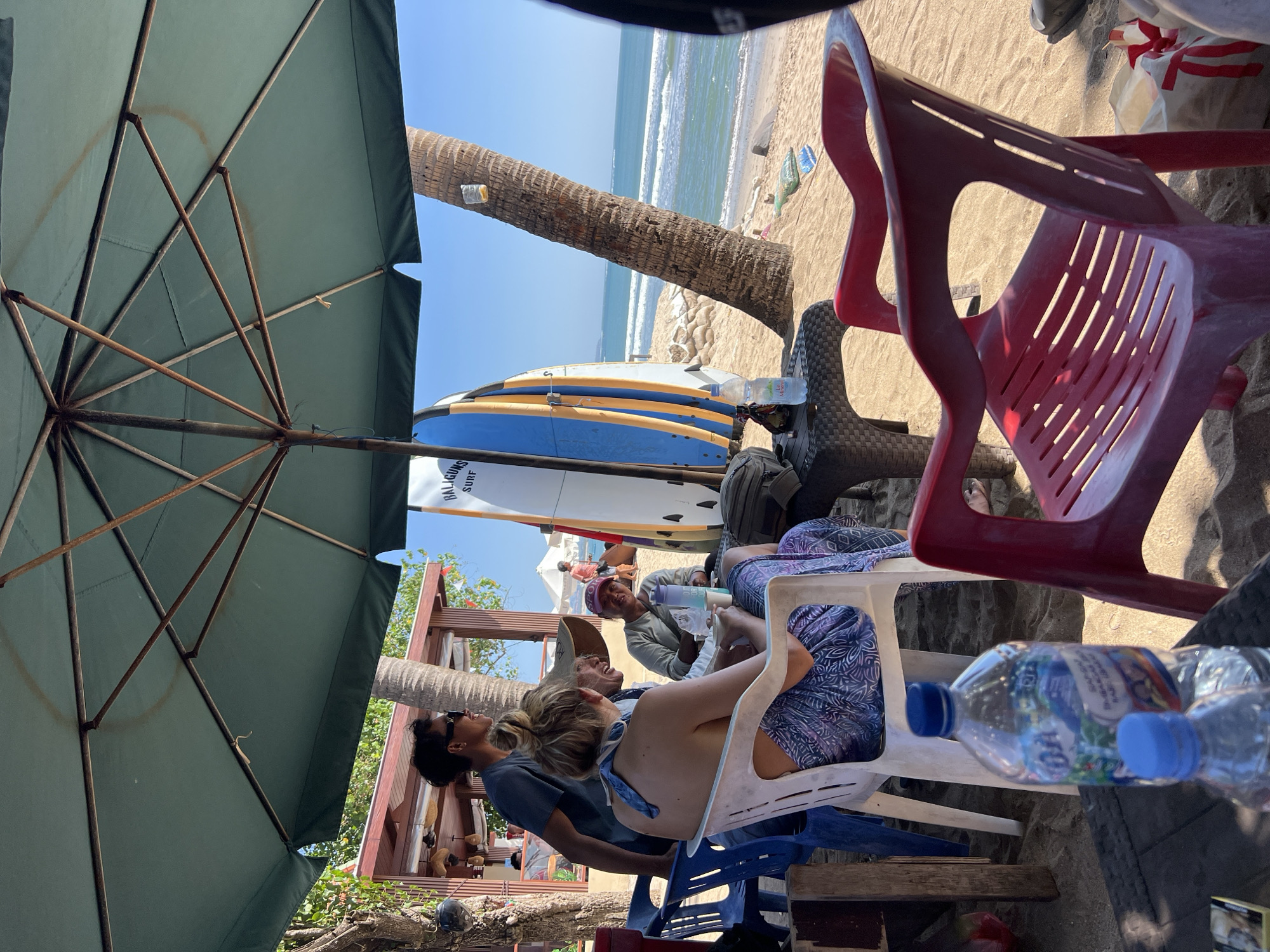 The image depicts a beach scene in Kuta, Indonesia. A group of people are seated under a large green umbrella, which provides shade from the sun. In the foreground, two plastic chairs, one red and one white, are visible on the sand. A table with a woven texture stands between the chairs, holding two bottles of water. Behind the table, a woman in a blue and white patterned skirt is seated in the white chair. A man wearing sunglasses and a blue t-shirt stands behind her. To their left, another woman with a hat and a light green shirt is seated.
In the background, a surfboard with blue and yellow accents leans against a palm tree. The ocean stretches out to the horizon, with gentle waves breaking on the sandy shore. The beach is dotted with palm trees and there are some structures, possibly shops or cafes, visible in the distance. The overall atmosphere appears relaxed, with people enjoying the beach environment.