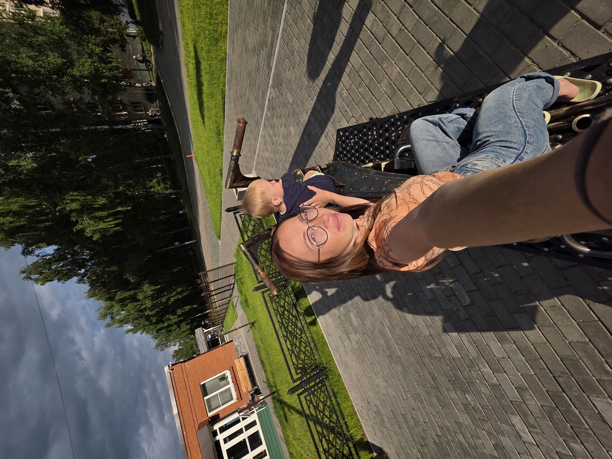 A woman with glasses and a young child are sitting on a playground ride resembling a train. The woman is taking a selfie, her arm extended towards the camera. She is wearing an orange patterned blouse and blue jeans. The child is wearing a dark blue shirt. They are on a paved area with a fence and green grass to their left. In the background, there is a brick building and trees. The image appears to have been taken in the city of Chaikovsky, Russia, as indicated by its likely presence within the city limits, given the architectural style and vegetation typical of the region. The lighting suggests it is late afternoon or early evening.