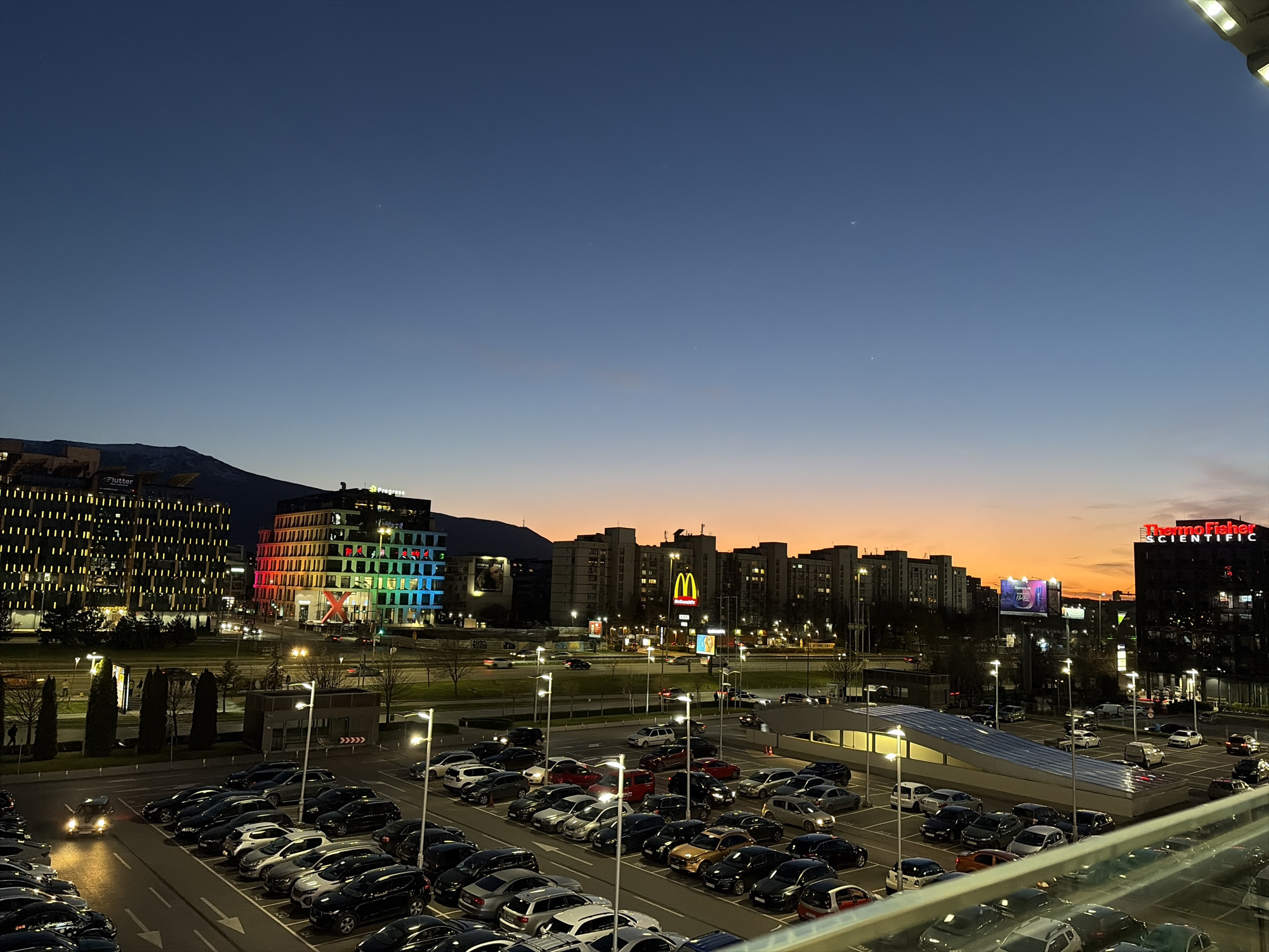 The image shows a dusk view of Sofia, Bulgaria, from an elevated position, likely a parking garage. In the foreground is a large, mostly full parking lot with numerous cars. Behind the parking lot, a roadway with moving cars is visible.
In the background, the cityscape includes several modern buildings. A prominent building is lit with rainbow colors, and another has a large red "X" on it. Other notable signs include a McDonald's golden arches and the "Thermo Fisher Scientific" sign.
In the far distance, a mountain range is visible against the fading light of the sunset. The sky is a gradient of blue to orange. No people are clearly visible.