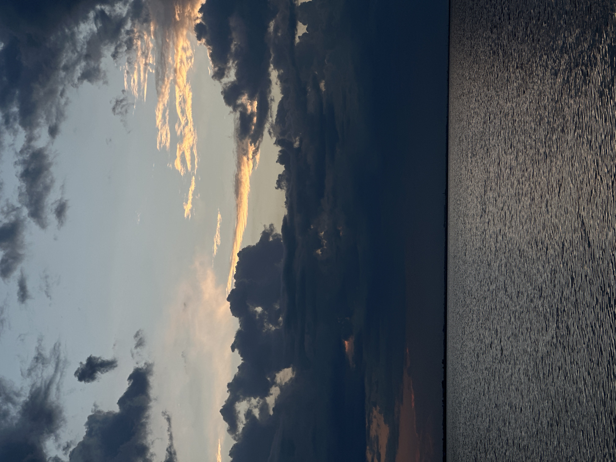 A wide shot captures a serene body of water under a dramatic, cloud-filled sky. The water, dark and rippling, fills the lower half of the frame, with a faint, dark horizon line separating it from the sky. The sky is a canvas of contrasting clouds, with large, dark, and imposing formations dominating the center and lower portions. Above them, lighter clouds are interspersed with streaks of warm, golden light, suggesting the presence of a setting or rising sun peeking through. The overall atmosphere is one of twilight or impending weather change. The scene is a natural landscape with no discernible human presence or urban structures, making it difficult to pinpoint a specific location within Kokshetau, Kazakhstan without additional context.