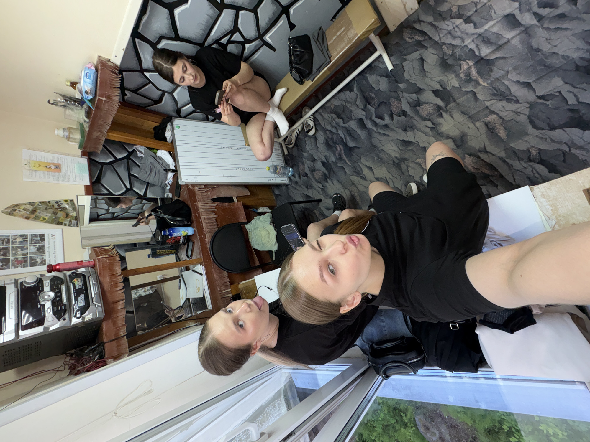 This media file captures three young women in what appears to be a casual indoor setting in Chaikovsky, Russia. Two women are in the foreground, taking a selfie. One, on the left, has her tongue playfully sticking out, while the other, on the right, poses with pursed lips. Both have dark hair pulled back and are wearing black tops; one is in jeans, and the other in black shorts.
In the background, a third woman is seated on a wooden bench against a wall with a distinctive painted gray and black stone-like pattern. She is wearing a black top and shorts with white socks, and appears to be looking at her phone.
The room has a varied decor. The floor is covered with a patterned carpet featuring shades of gray and brown. A large window on the right reveals an overcast sky and green foliage outside, indicating it's daytime. A stereo system and other various items are visible on shelves and surfaces. On a white board in the background, Cyrillic text is visible, including "ЧАЙКОВСКИЙ" (Chaikovsky), confirming the location, and "ПРИВЕТ" (Privet, meaning Hello). Other objects include a large brown broom, various bottles, and bags. The overall atmosphere is relaxed and informal.