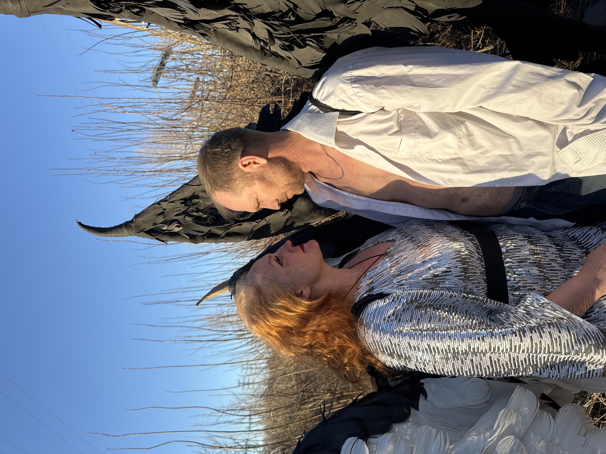 A woman and a man are lying down outdoors with what appears to be wings attached. The woman has red hair and is wearing a shiny, silver, sequined top. The man is wearing an unbuttoned white dress shirt. They are lying head-to-head, facing each other. The wings appear to be black on one side and white on the other. The background includes leafless trees and a blue sky. The location is Chaikovsky, Russia.
