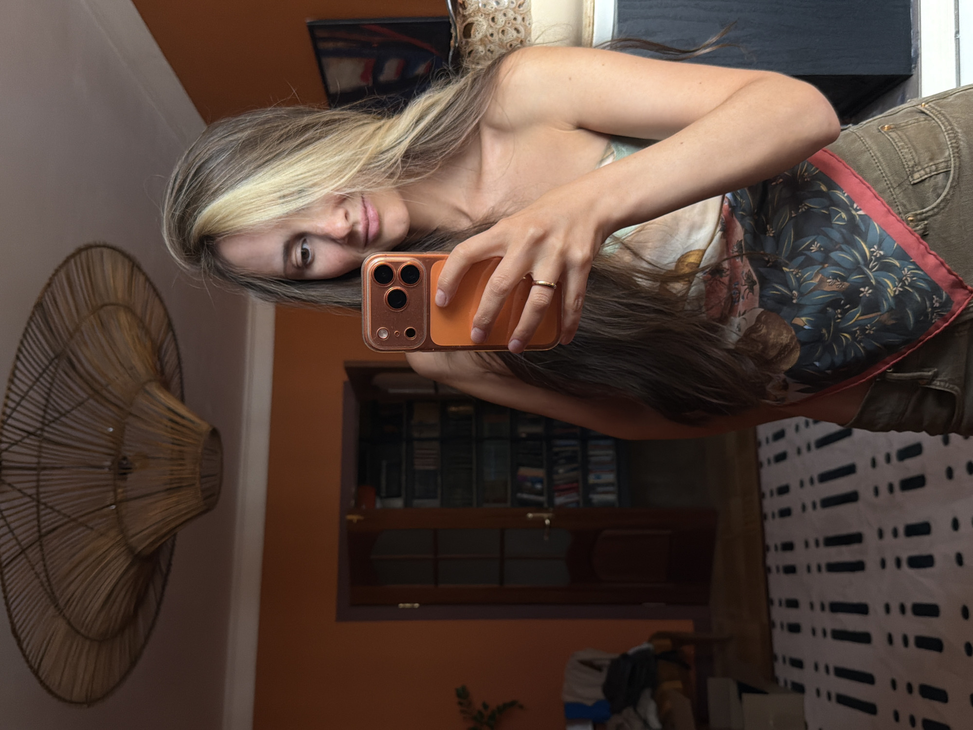 This media file is a vertical, full-body, mirror selfie taken indoors. The subject is a woman with long, wavy brown and blonde hair, taking the photo with her phone. She is wearing a patterned top and khaki pants. Her left arm is bent, and her right hand holds the phone. She is wearing a ring on her right ring finger. The background shows a room with an orange wall, a dark cabinet, a wooden cabinet with shelves, and a patterned tapestry or rug on the floor. A large, wicker decorative fan is mounted on the wall to the left. The lighting appears to be natural, suggesting daytime. The overall mood is casual and relaxed. There is no visible text in the image. The location context is Khimki, Russia.