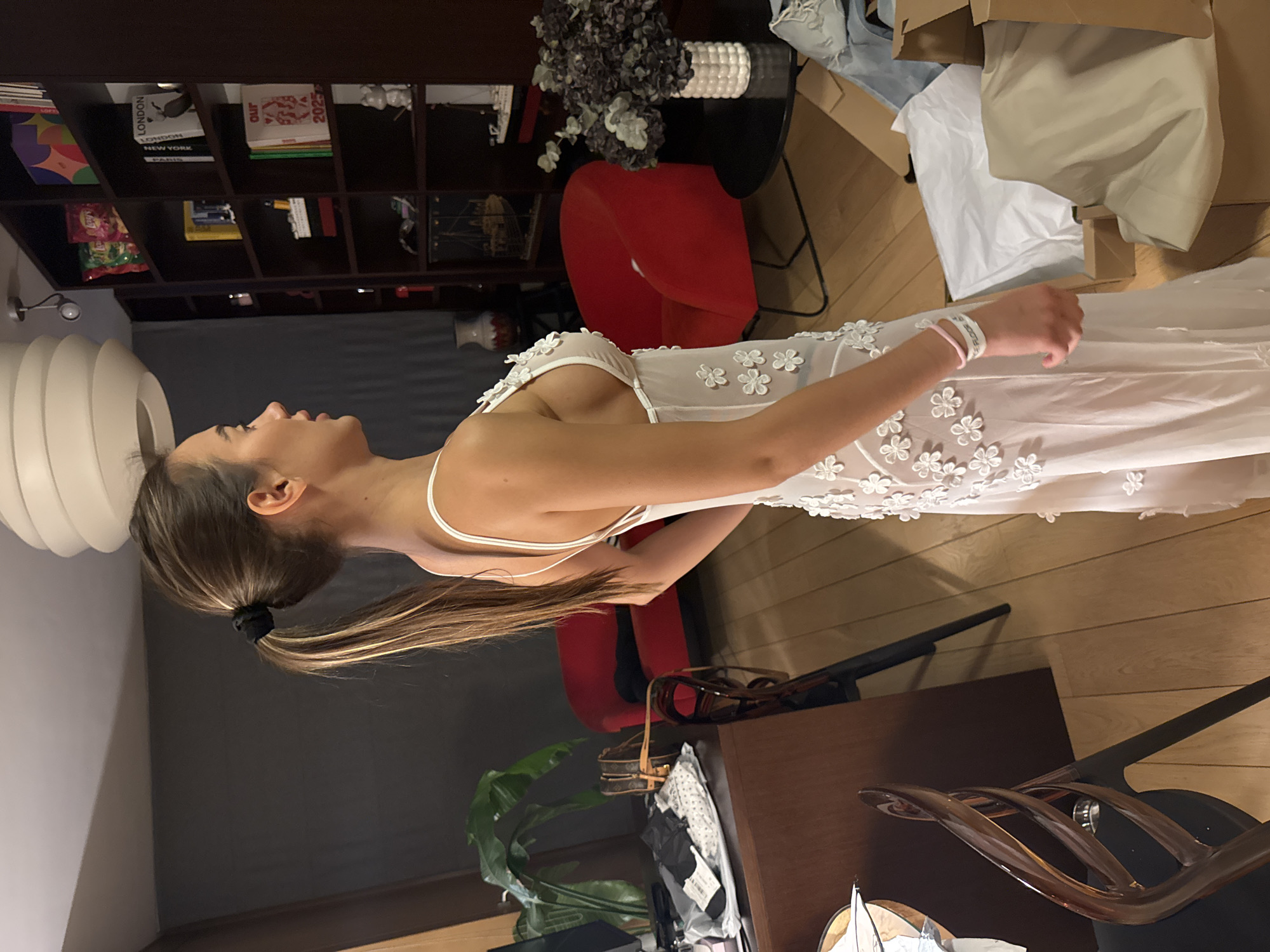 A woman with light brown hair in a ponytail is standing indoors, wearing a white, sheer dress with floral appliqués. She is positioned in a room with dark grey walls and light-colored hardwood floors. Behind her, there is a bookshelf filled with books and decorative objects. To her right, there is a red chair and a black side table with a vase of flowers. In the foreground, there is a brown desk with various objects on it, including a purse and papers. The overall setting suggests a residential or apartment interior. While the location is stated as Warsaw, Poland, there is nothing in the image to definitively confirm this.