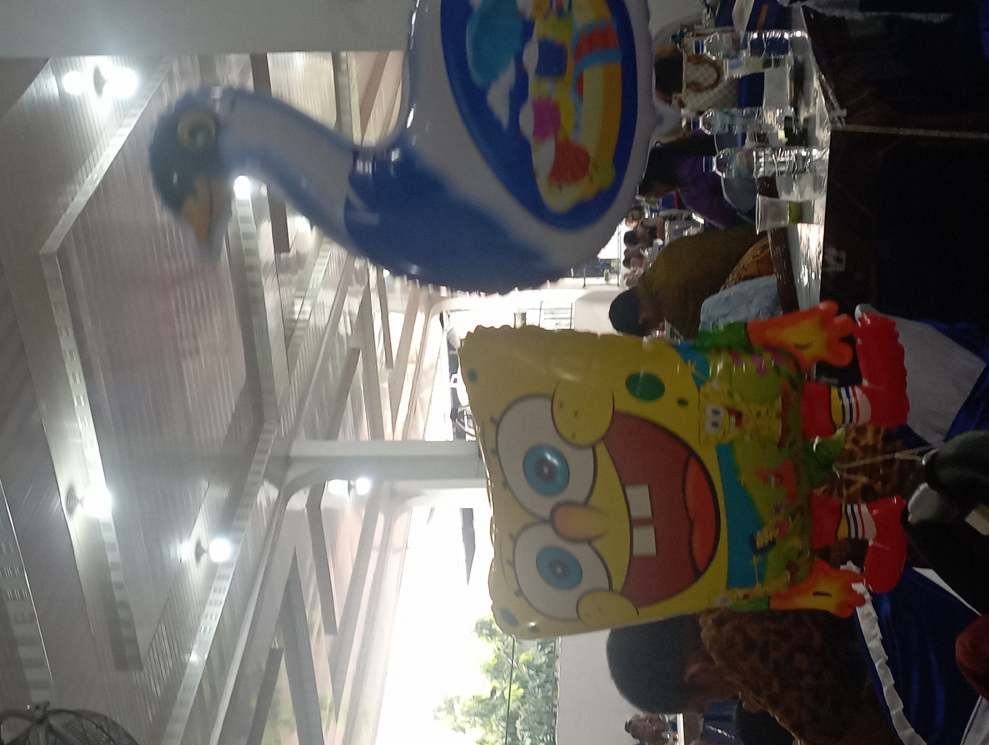 An indoor scene, likely a reception hall, is depicted with a Spongebob Squarepants balloon and a large blue and white swan-shaped balloon prominently displayed. The Spongebob balloon, in the foreground, features the character's signature yellow face, large eyes, and wide smile. The swan balloon, to the right, has a blue body with white accents and an illustration of a scene with ice cream cones on its side.
The background reveals a ceiling with exposed beams and fluorescent lighting. Tables are set up, with several people seated and engaged in conversation or dining. Water bottles and plastic cups are visible on the tables. The overall ambiance suggests a casual gathering or celebration. The environment appears to be a temporary setup, possibly for an event.