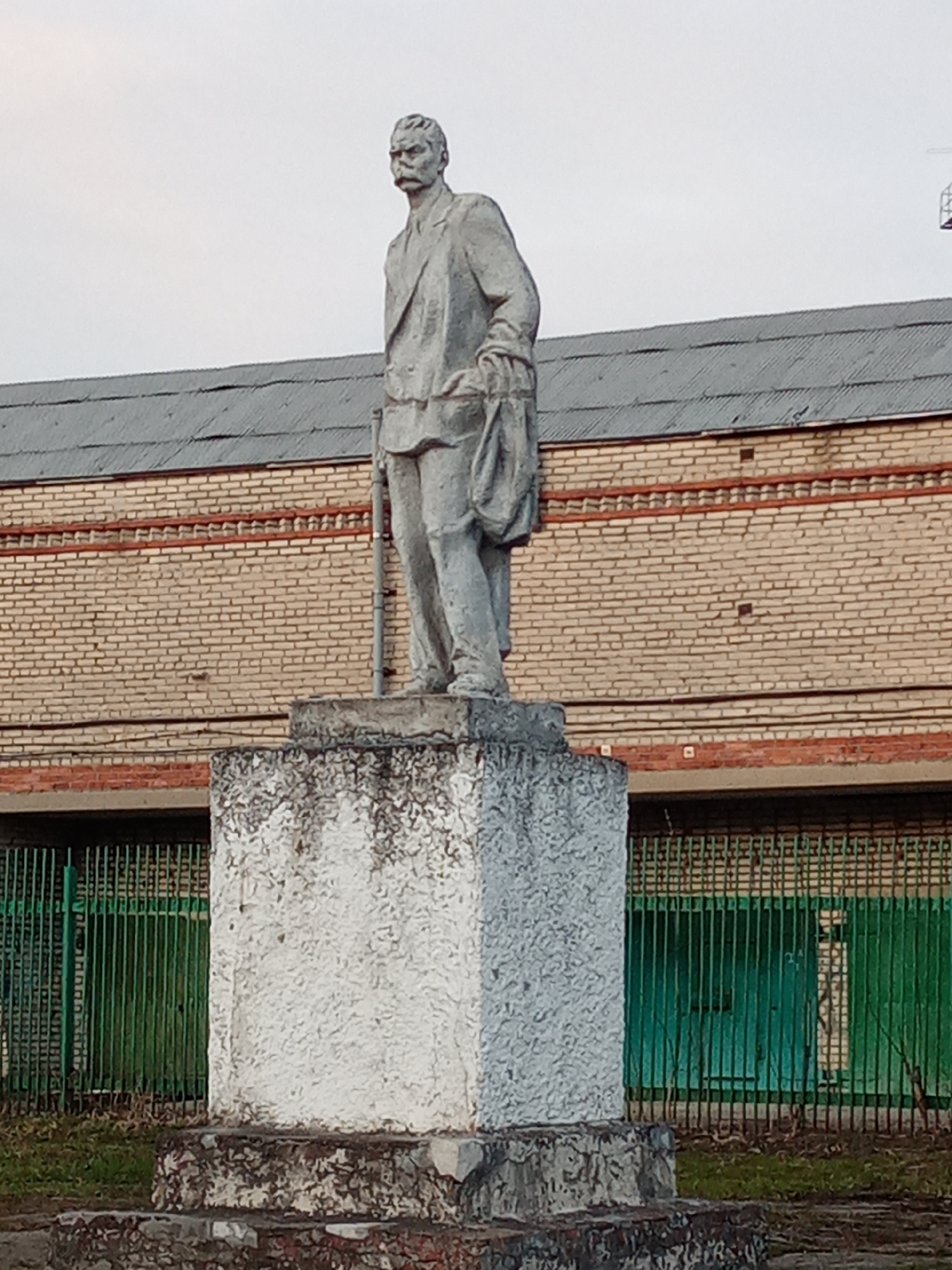 The image depicts a weathered statue of a man, likely a historical or political figure, standing on a concrete pedestal. The statue appears to be made of stone or concrete and shows signs of age and environmental exposure. The man is dressed in a suit and has a stern expression.
The background includes a brick building with a corrugated metal roof and a green metal fence. The overall environment appears to be an urban setting, possibly a public space or a park in Elektrostal, Russia.
