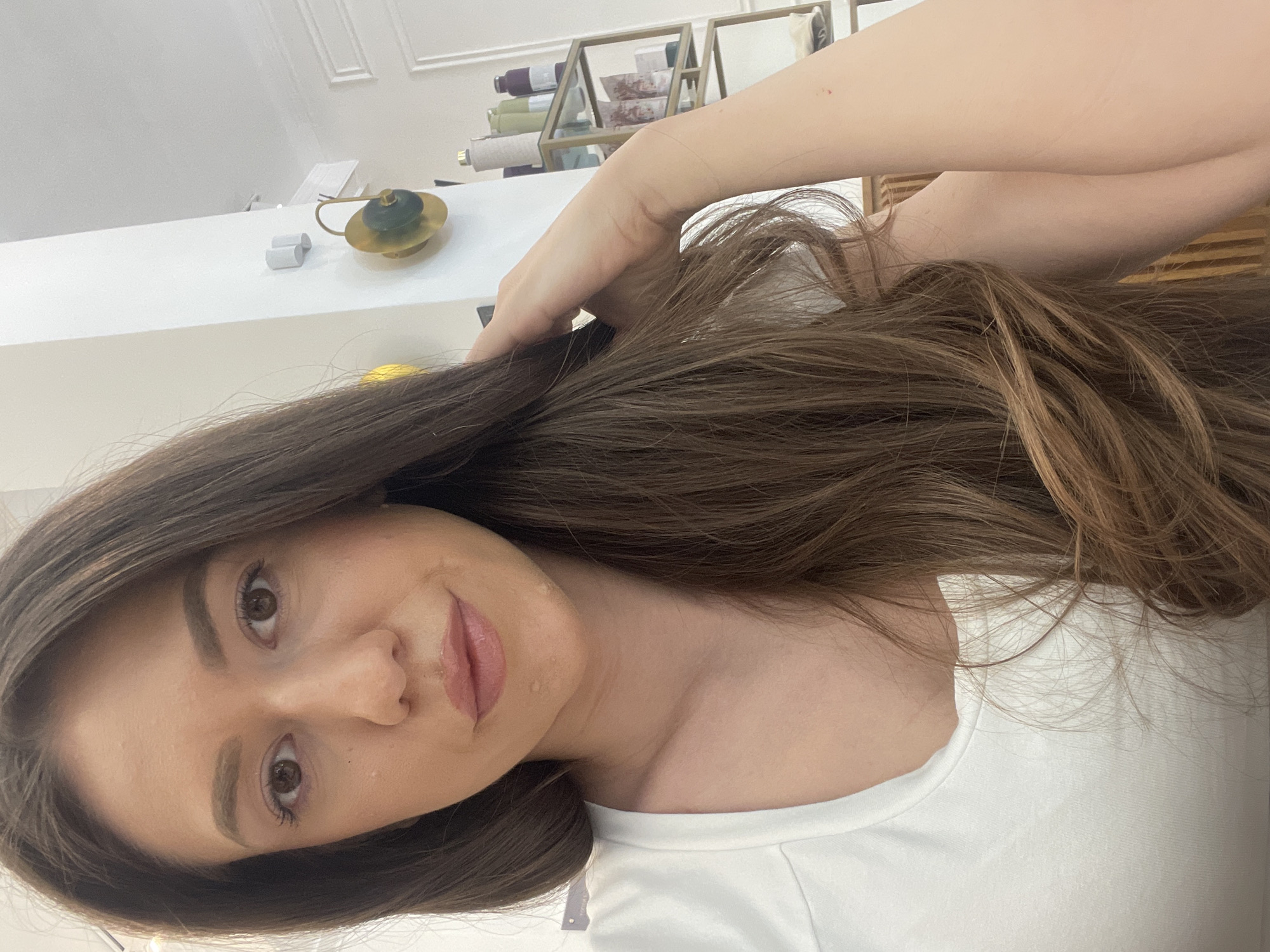 The image shows a young woman with long, brown hair, light skin, and brown eyes, wearing a white T-shirt. She is likely taking a selfie, as she is holding her hair with one hand. The background includes a white counter or shelf, some decorative items in a gold-colored display case, and parts of a wall with molding. While it is impossible to determine the location as Sofia, Bulgaria, from this image alone, the style of the furnishings and the woman's appearance suggest an interior setting.
