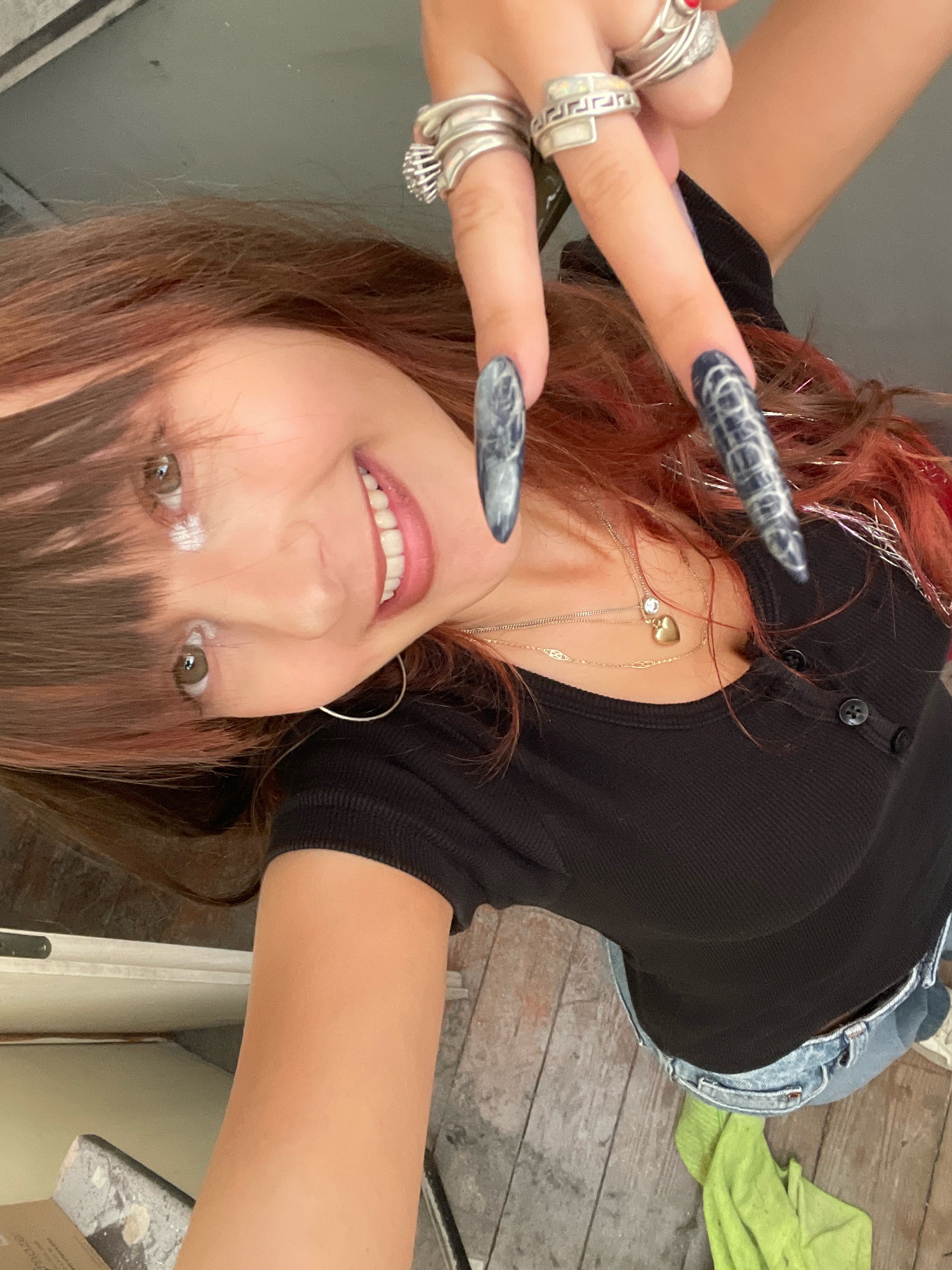 The image shows a close-up, tilted selfie of a young woman with long, reddish-brown hair and bangs. She is wearing a black, ribbed, short-sleeved shirt and blue jeans. She has long, dark nails with a marbled pattern and is wearing several silver rings on her fingers, including one with a Greek key design. She is also wearing a delicate gold necklace with a heart pendant and a small blue stone. The woman is smiling, showing her teeth, and has a smudge of white makeup on her cheekbone. Her eyes are brown and she appears to be indoors, possibly in a home or studio. The background includes a wooden floor and part of a white object, possibly furniture or a wall. There is no indication of the specific location within Raanana, Israel.
