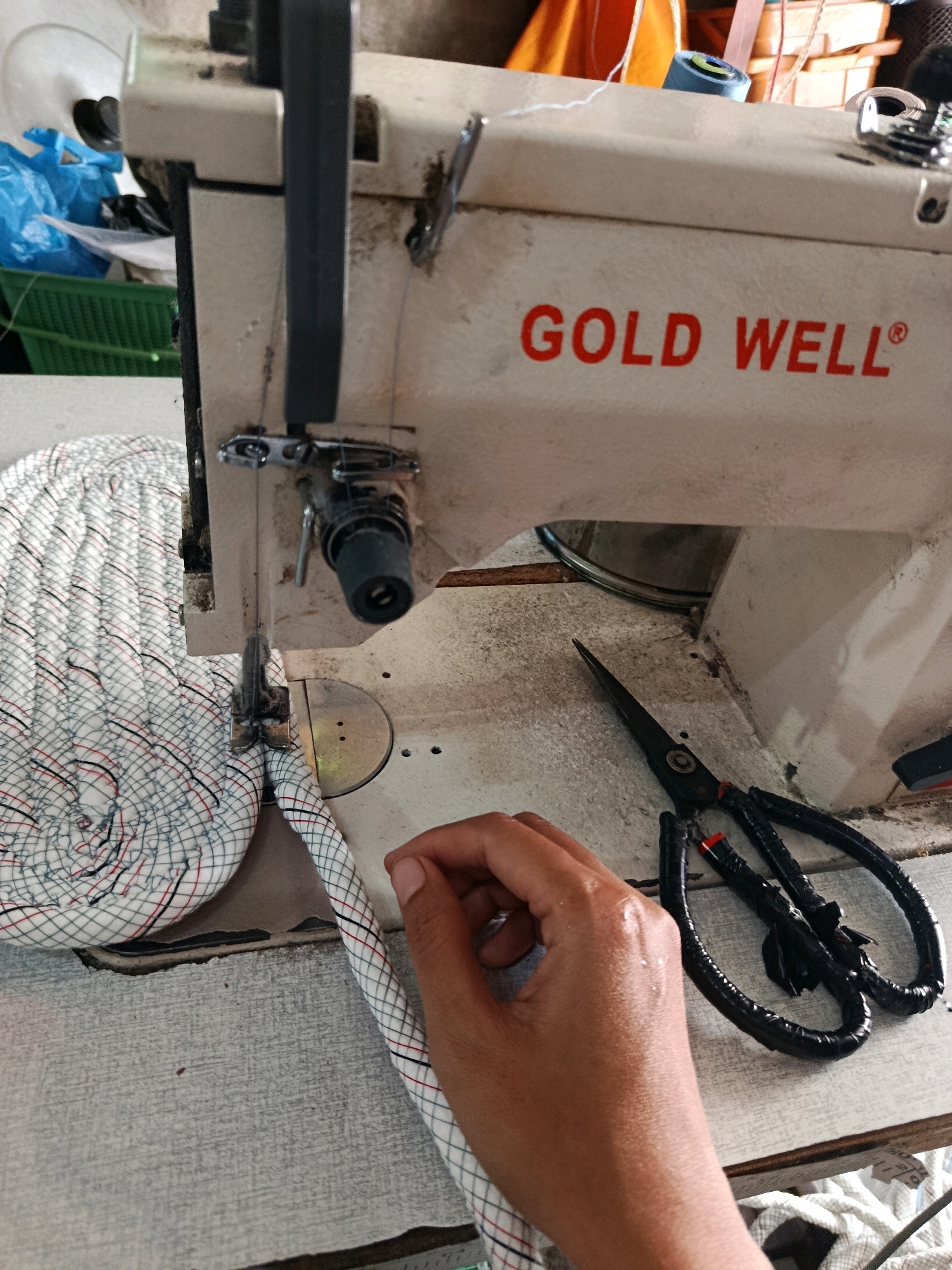 A close-up, eye-level shot shows a Gold Well sewing machine in what appears to be a workshop or home-based sewing area in Karang Rejo, Indonesia. The machine is beige with red lettering on the front. A coil of white, red, and black checkered fabric rope is on the left, with a section of the rope being fed into the sewing machine's needle. A person's hand, with a slightly wet appearance, is positioned to guide the rope into the machine. A pair of black-handled scissors rests on the machine's baseplate. In the background, partially visible, are blue plastic bags, a green basket, and some orange fabric. The overall environment suggests a practical, hands-on work setting.
