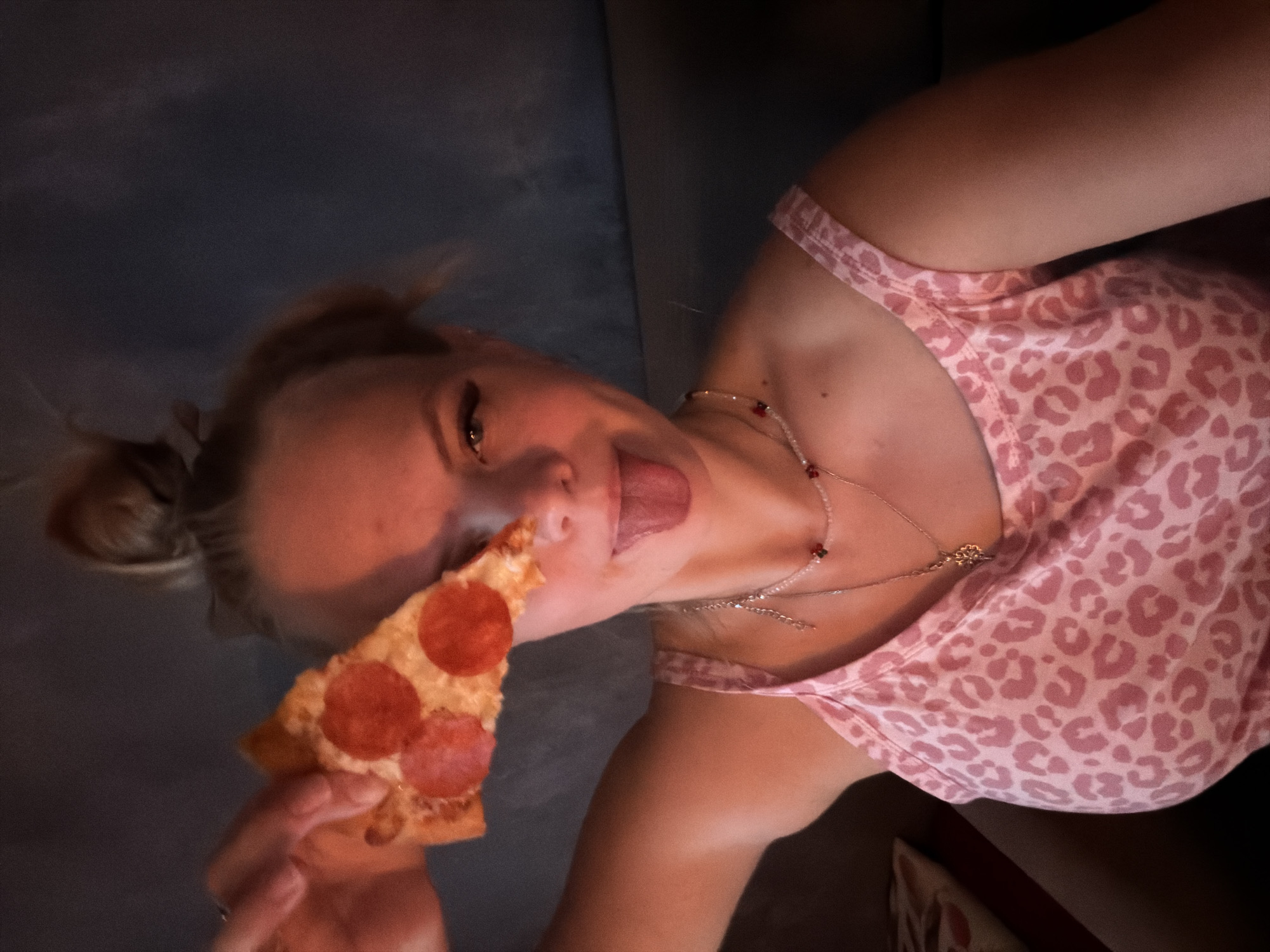 The image shows a young woman holding a slice of pepperoni pizza up to her face, obscuring her right eye. She is sticking her tongue out and looking towards the camera. She is wearing a pink tank top with a leopard print pattern and several necklaces. The background is dark and out of focus, suggesting an indoor setting. There is no visible indication of the specific location within Chaikovsky, Russia.