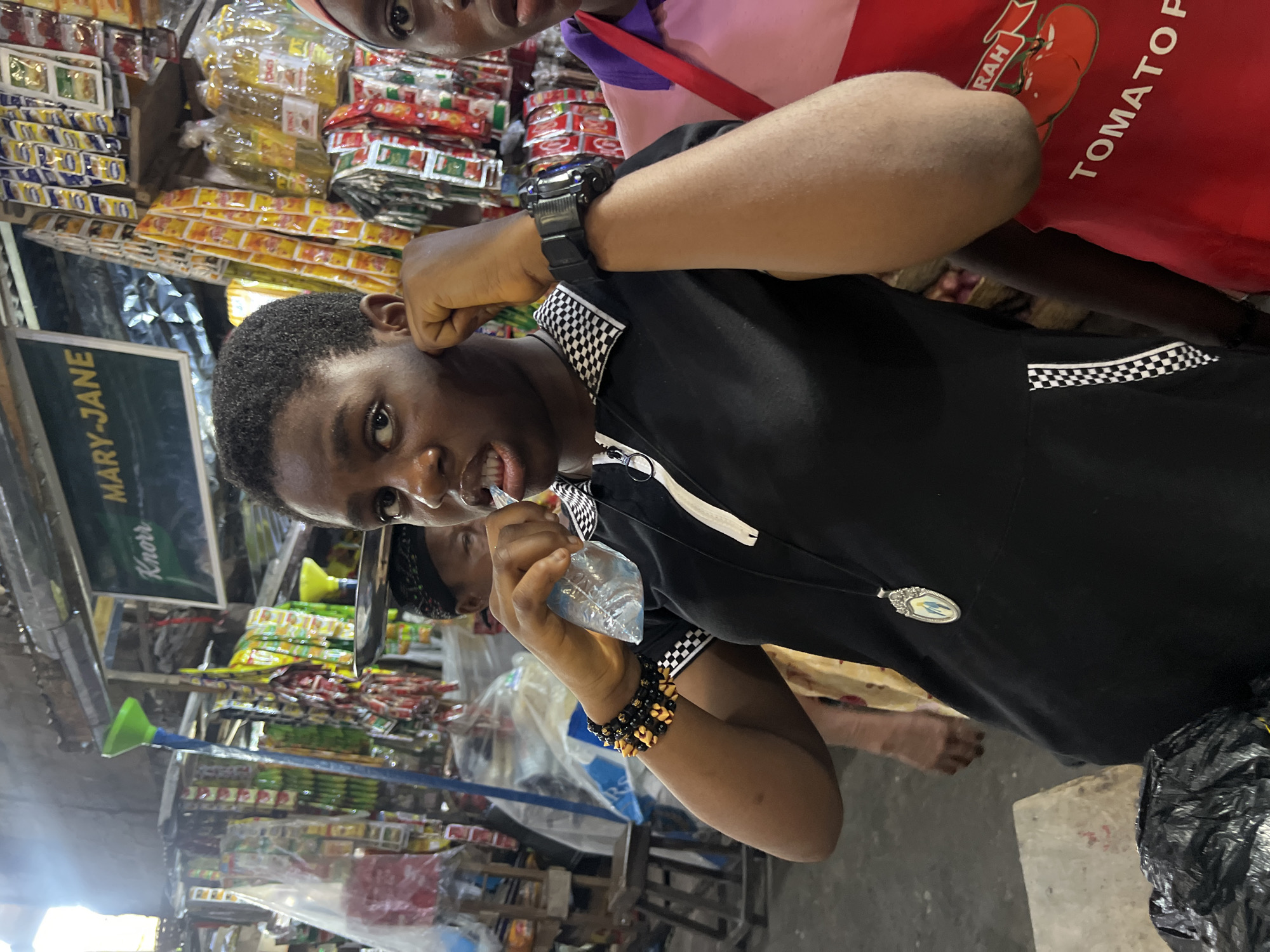 A medium shot captures a young woman in a black zip-up shirt with a checkered collar, standing in a market in Nkpor, Nigeria. Her dark hair is short, and she is holding a small plastic bag of water to her mouth while touching her ear with her right hand. She wears beaded bracelets on her left wrist and a silver pendant necklace. A person to her right, wearing a pink shirt and a red apron with "TOMATO P..." visible, is partially visible. The background is filled with shelves of packaged goods, including Knorr products and other food items. The market appears to be outdoors or in a covered open-air space, with natural light filtering in from the left. A wooden stool is visible to the left of the frame.