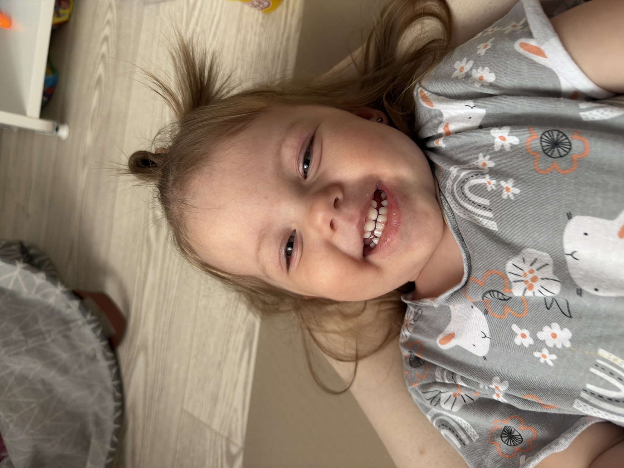 A young child with light brown hair and a small ponytail on top of their head is lying on their back. The child is smiling and wearing a gray shirt with a pattern of white rabbits, flowers, and rainbows. The background consists of a light-colored wooden floor, the edge of a white cabinet, and a gray patterned object in the lower left corner. The location in the city of Reshetnikovo, Russia is unknown.