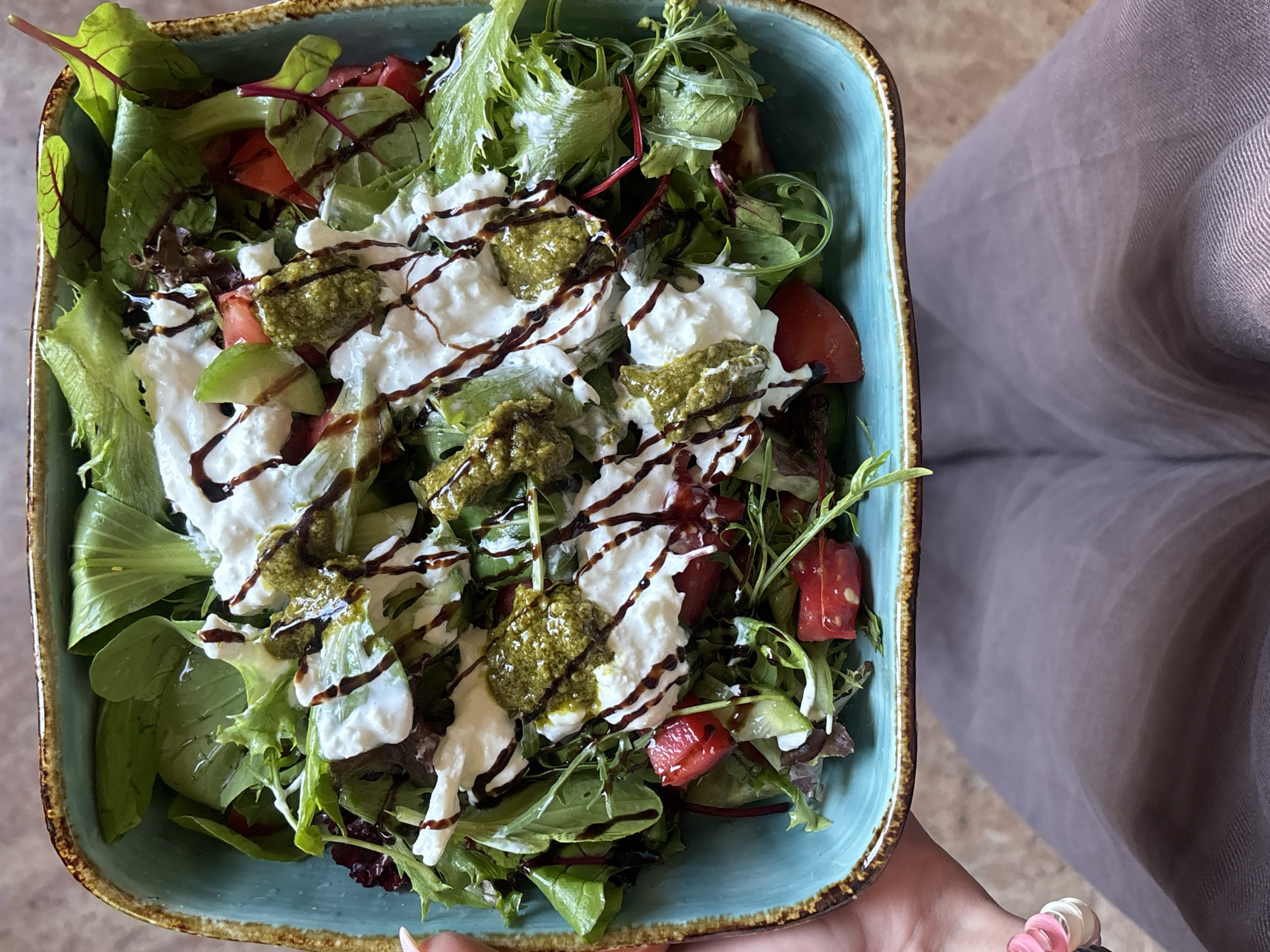 A person holds a blue square bowl filled with a salad. The salad includes lettuce, tomatoes, white cheese (possibly burrata), pesto, and balsamic glaze. The person is wearing grey pants and has a pink and white bracelet. The location is unknown; there is no indication it is Yerevan, Armenia.