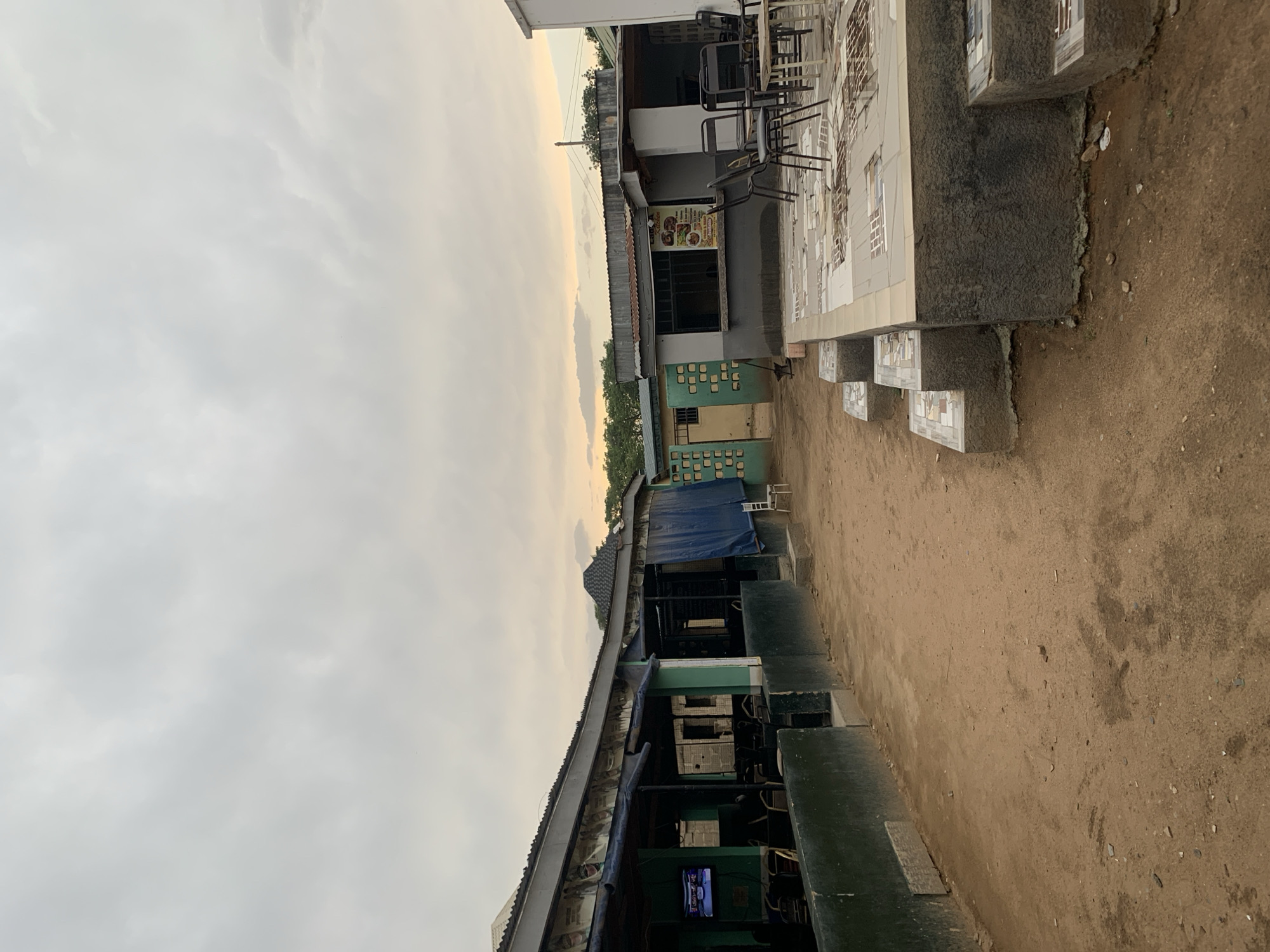 The image depicts an outdoor scene in New Stadium, Nigeria, characterized by informal commercial structures along a dirt road. The sky is overcast, suggesting a cloudy day. Several single-story buildings are present. One building features a partially tiled façade with visible seating arrangements and tables, suggesting a restaurant or eatery. Another structure, possibly a shop or stall, has a blue tarpaulin roof. The general environment appears to be somewhat dilapidated, with unpaved ground and simple building materials. There are no people visible in the shot.
