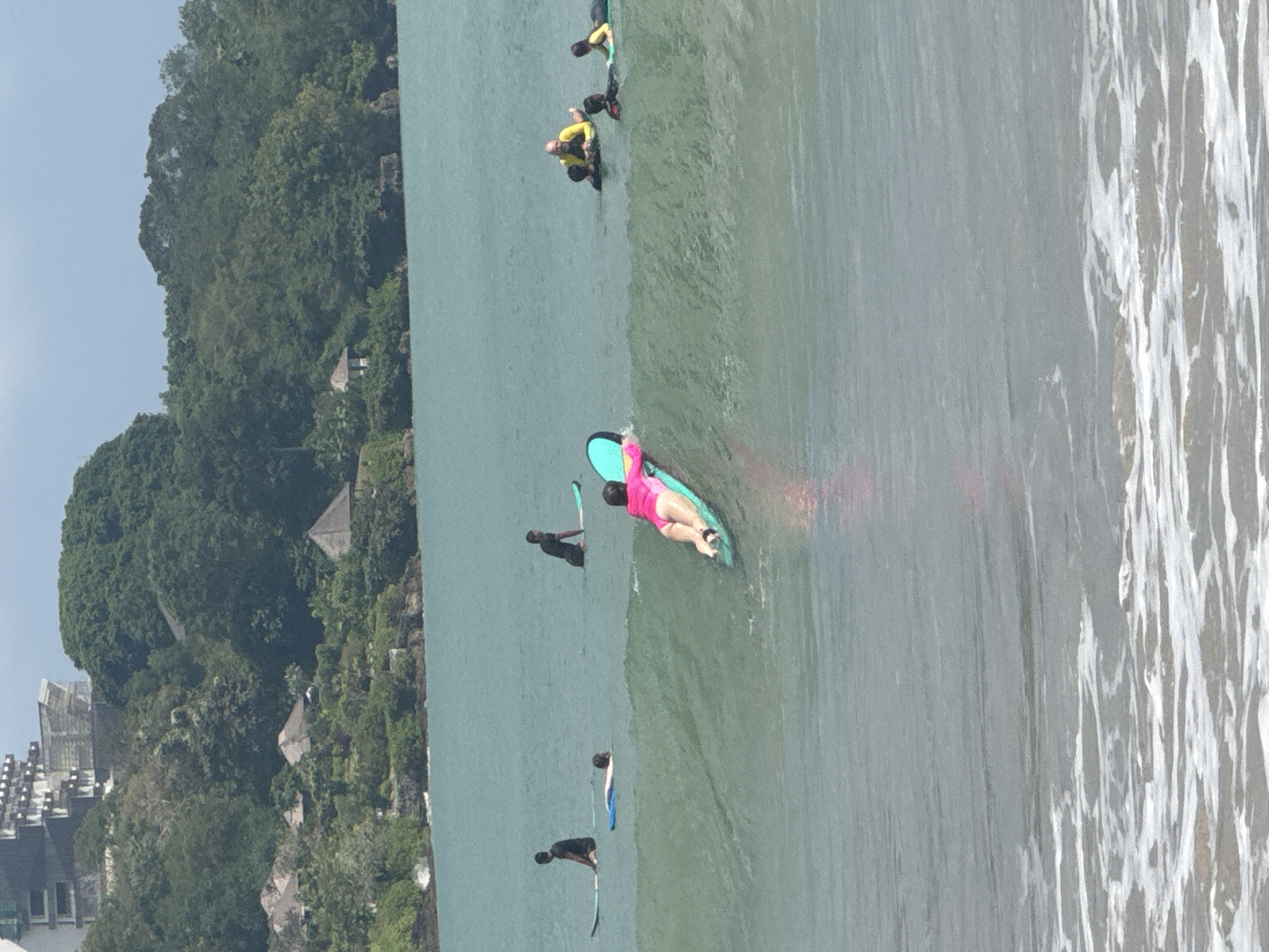 The image presents an overhead view of a coastal scene in Kuta Selatan, Indonesia, featuring several individuals engaged in water sports. The main subject in the foreground is a person wearing a bright pink swimsuit, lying prone on a turquoise and blue surfboard, seemingly paddling or waiting for a wave.
Further back in the calm, greenish-blue water, other people are visible with their boards. One person in dark attire stands on a dark surfboard or paddleboard, holding a paddle. Another individual is seen lying on a surfboard. In the mid-ground, a group of people, some wearing yellow rashguards, are also on surfboards. The water transitions from relatively calm to a band of white foam and gentle waves on the right side of the frame.
The left side of the image is dominated by a verdant, densely vegetated hillside or cliff, covered in lush tropical trees and plants. Scattered amongst the greenery are several buildings or huts with dark roofs, blending into the natural landscape. In the distant background on the far left, a more developed area with multi-story buildings is partially visible.
The scene unfolds during daylight hours, characterized by bright, even lighting, suggesting clear weather. The overall impression is one of leisure and outdoor recreation in a tropical, coastal environment. There is no visible text in the media.